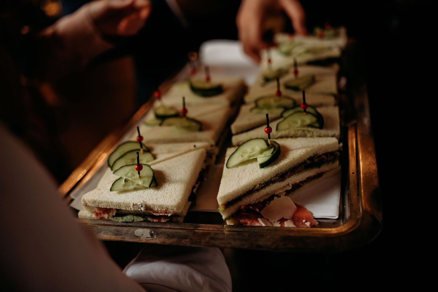 Elegant finger sandwiches with cucumber garnish served on a silver tray at a wedding reception.