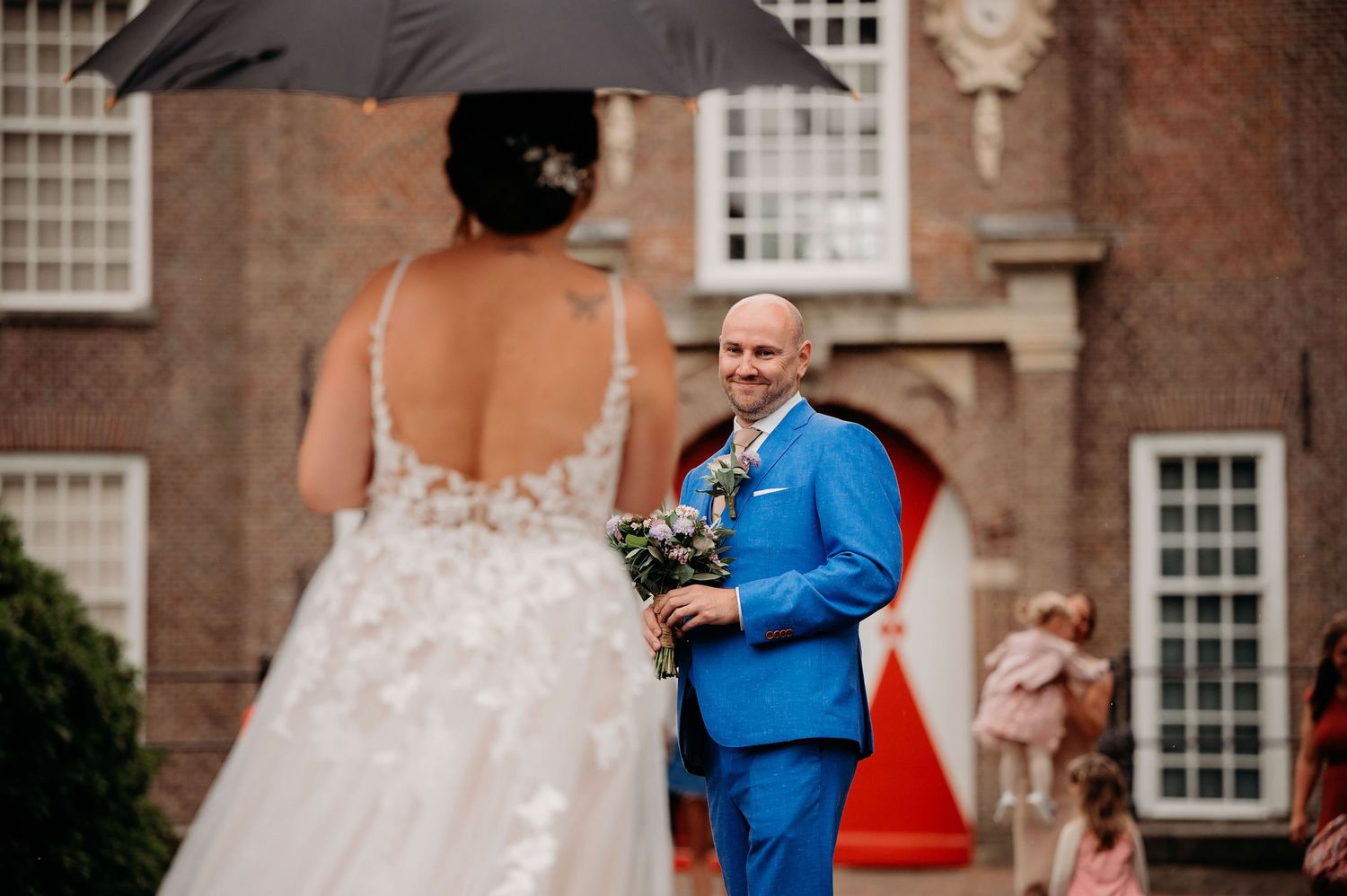 Groom in blue suit smiles at bride in lace wedding dress during outdoor first look at brick manor.