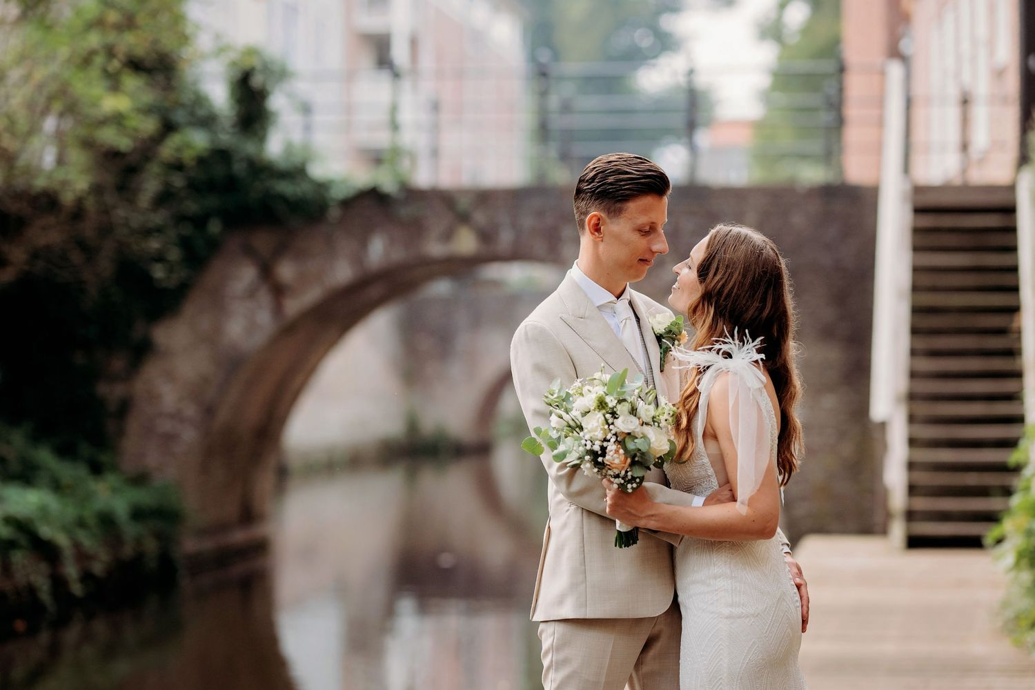 Bride and groom sharing a romantic moment by a canal bridge, holding a white bouquet on their wedding day.