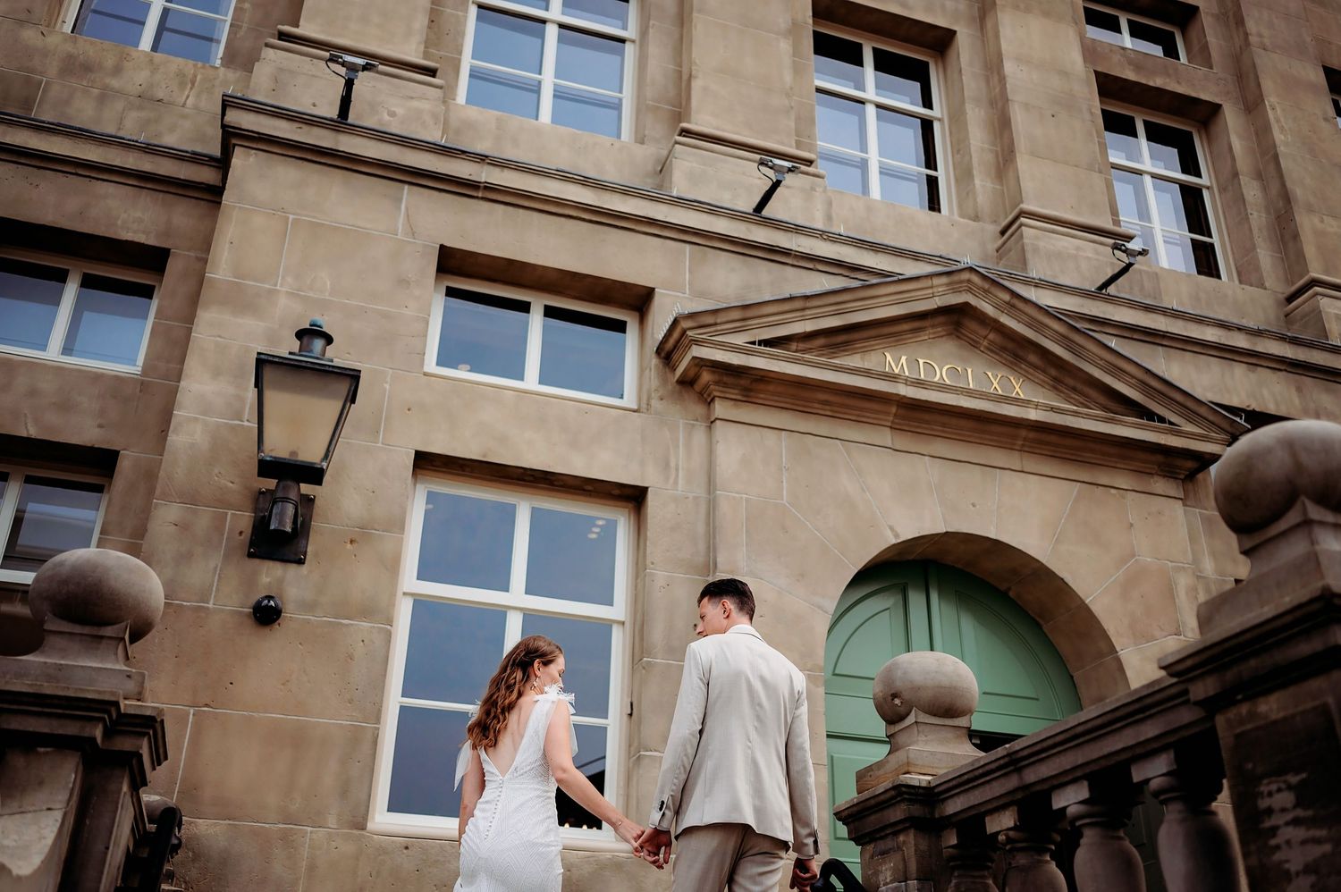Couple holding hands on stone steps outside a grand historic building with green doors on their wedding day.