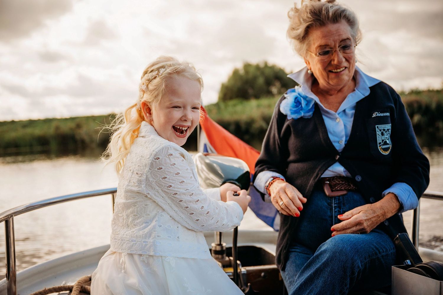 Young girl in white dress laughs joyfully with elderly woman on a boat on a calm river.