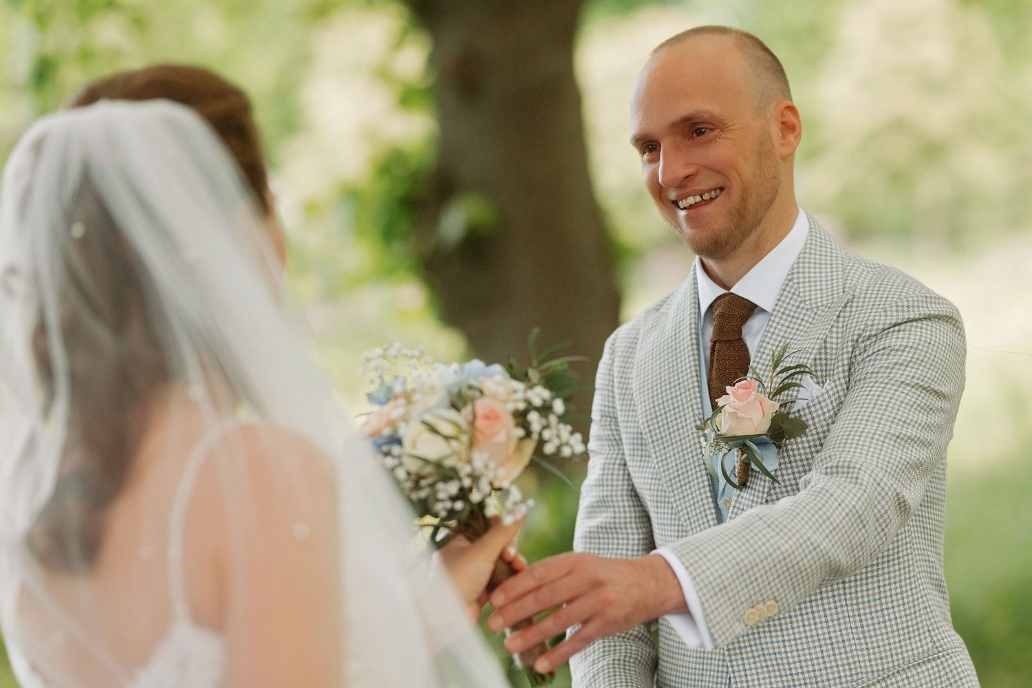 Groom in light gray suit smiling at bride during outdoor wedding ceremony with floral bouquet.