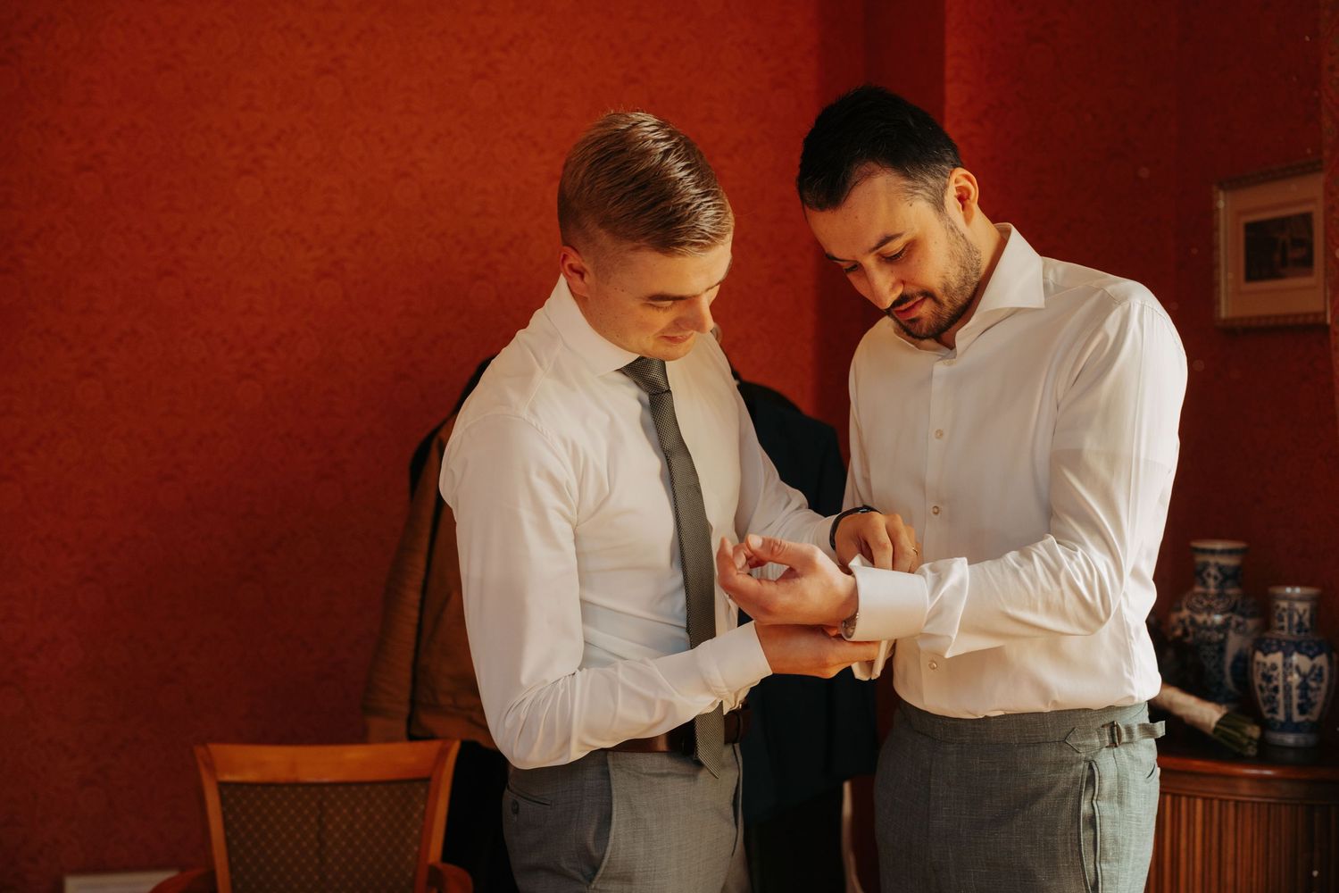 Two men in white shirts stand against a red wall as one helps the other fasten his cufflink while getting ready for a wedding.