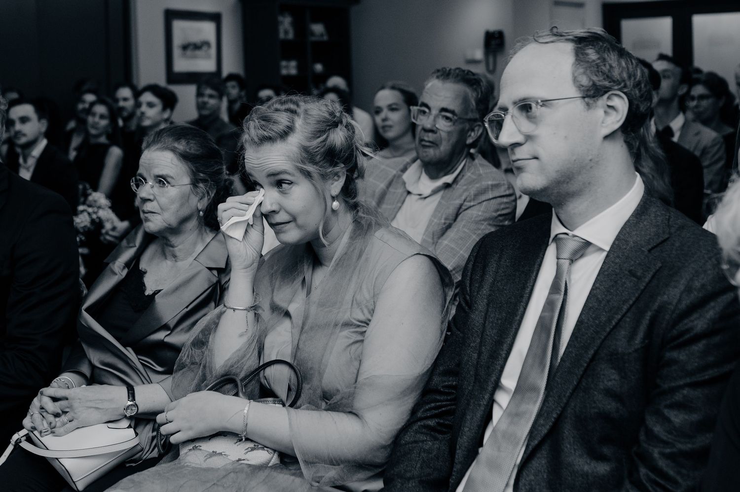 Black and white photo of audience members at a formal event, with a woman wiping tears and a man in glasses beside her.