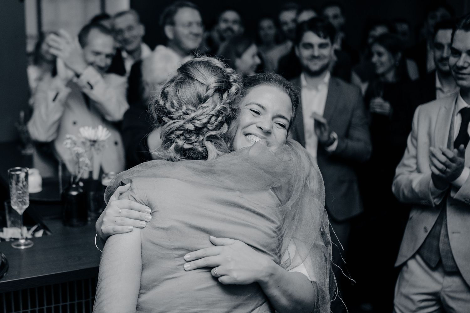 Bride shares joyful hug with smiling guest at wedding reception in black and white.