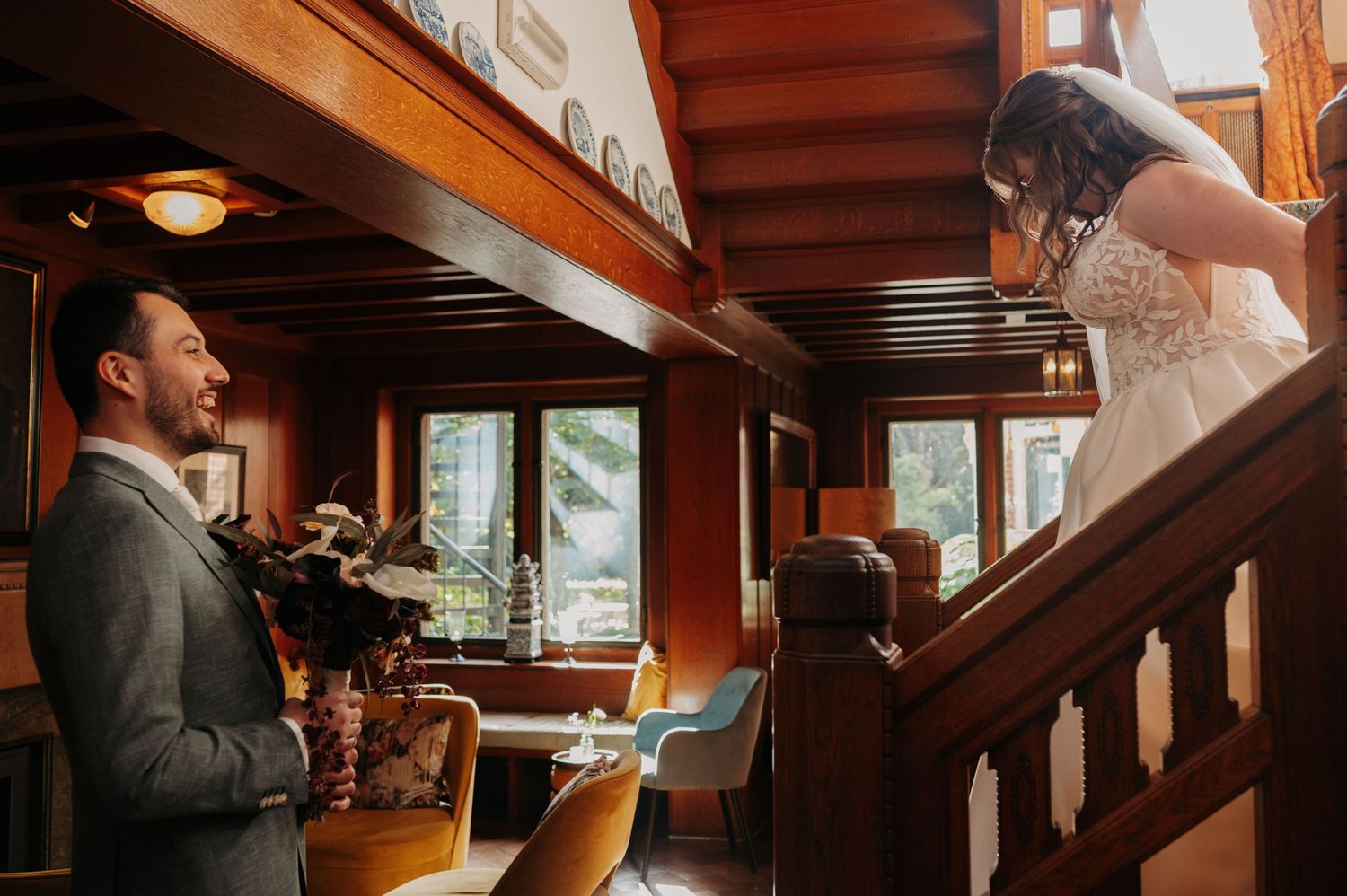 Groom in a gray suit looks up at a bride in a lace gown on a wooden staircase inside a rustic lodge with large windows.