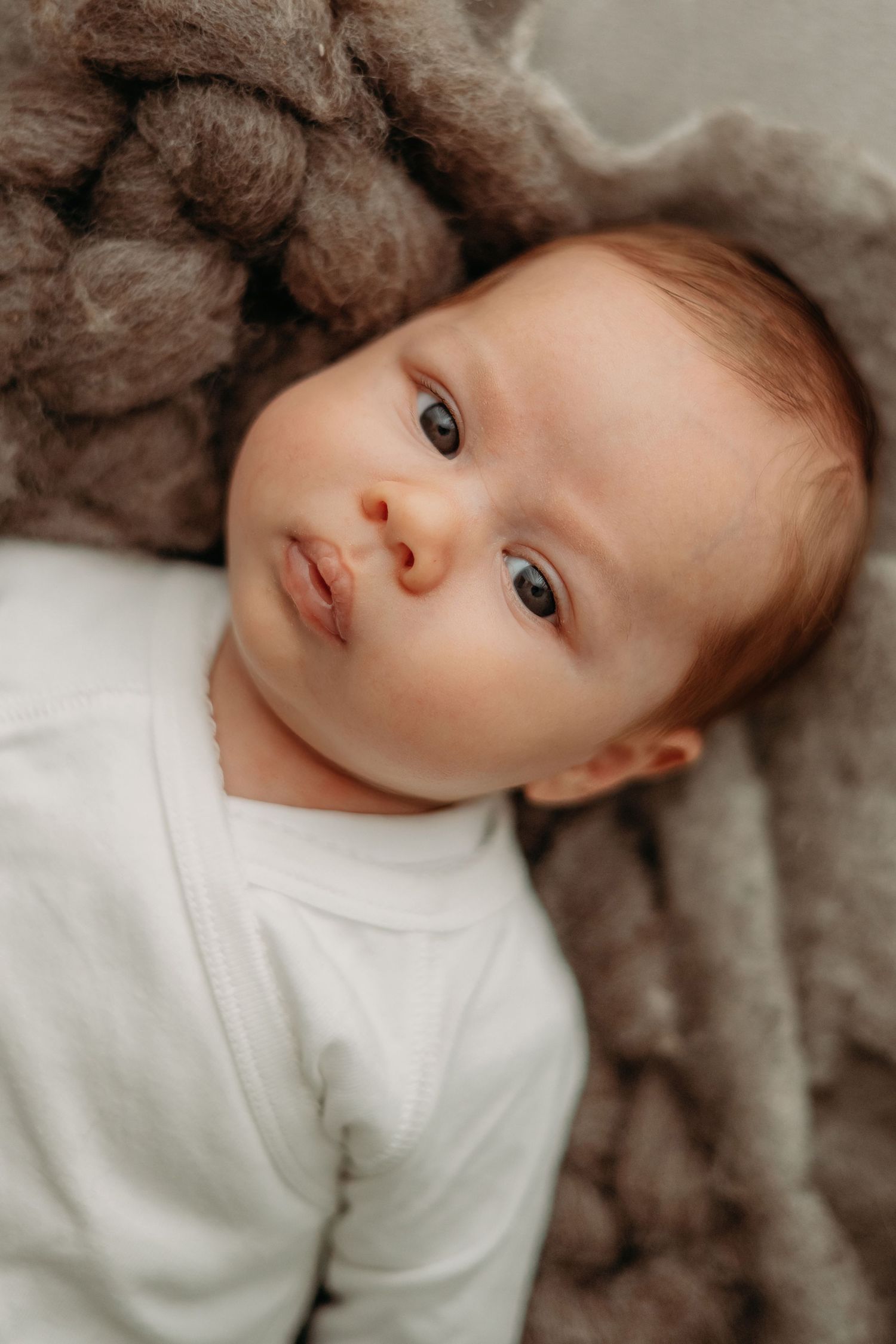 Baby in witte outfit liggend op zachte grijze deken met nieuwsgierige blik, newbornfotografie Brabant