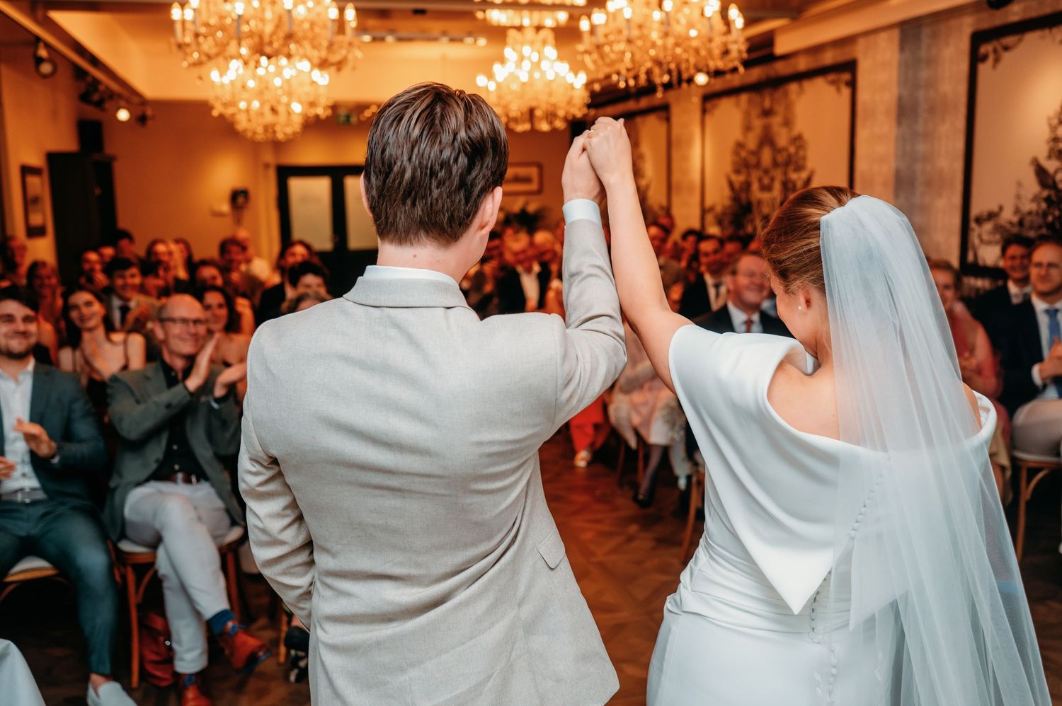 Newlyweds raise joined hands triumphantly before applauding wedding guests in a chandelier-lit reception hall.