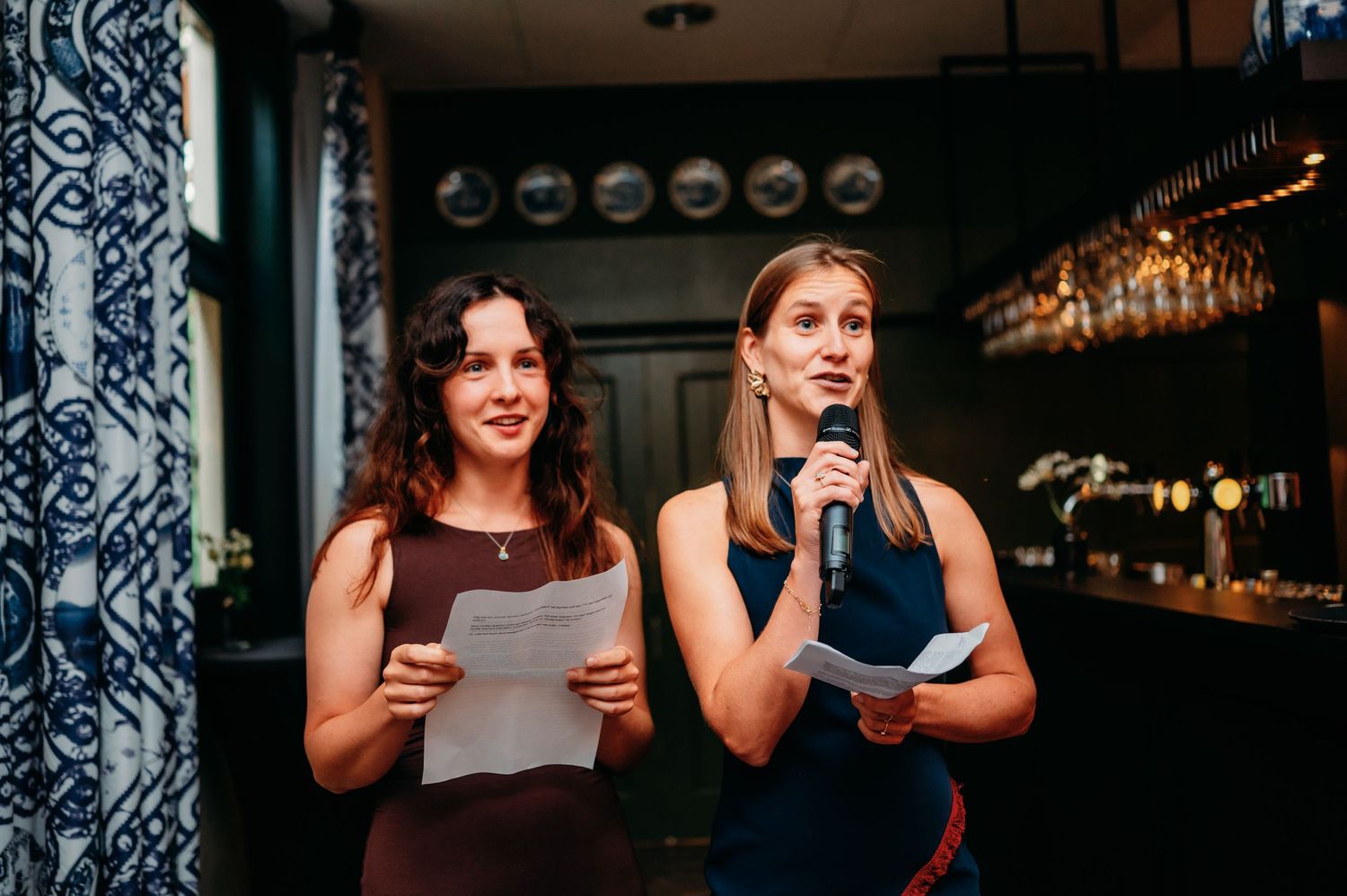 Two women in a decorated bar venue deliver a wedding speech together while holding papers and a microphone.