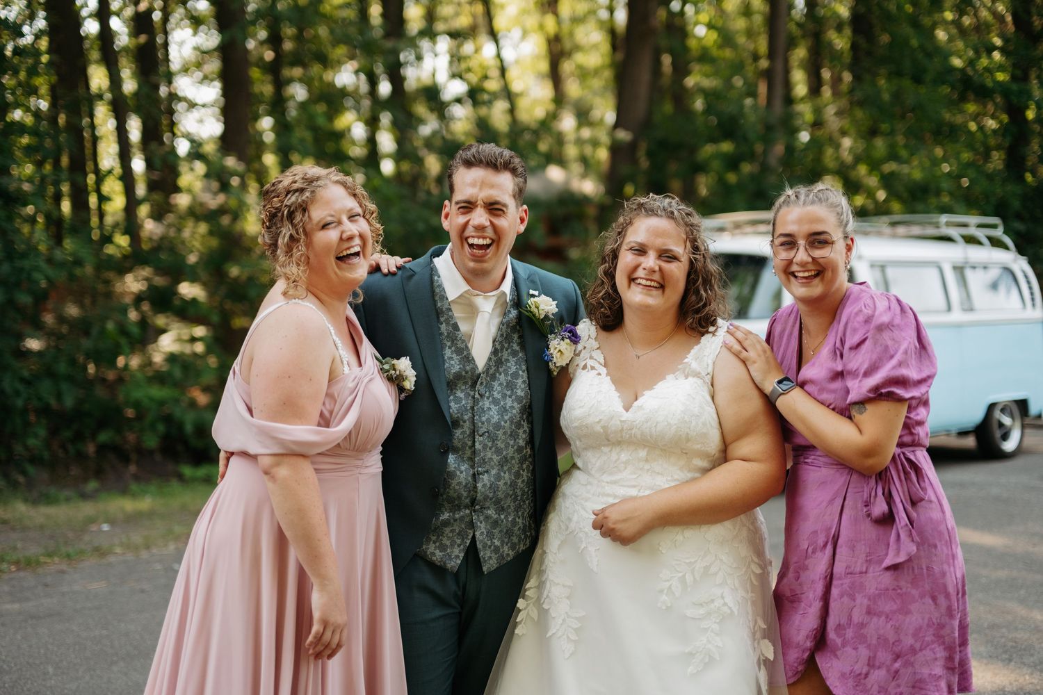 Joyful wedding group photo with bride, groom, and two guests laughing outdoors near a vintage blue van.