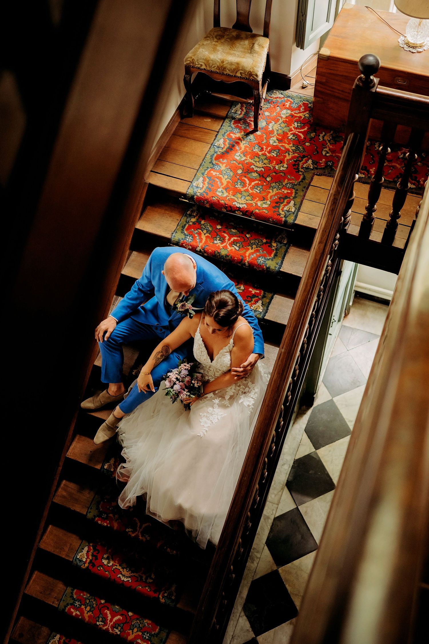 Aerial view of bride and groom sharing a romantic moment on ornate wooden staircase with red carpet runner.