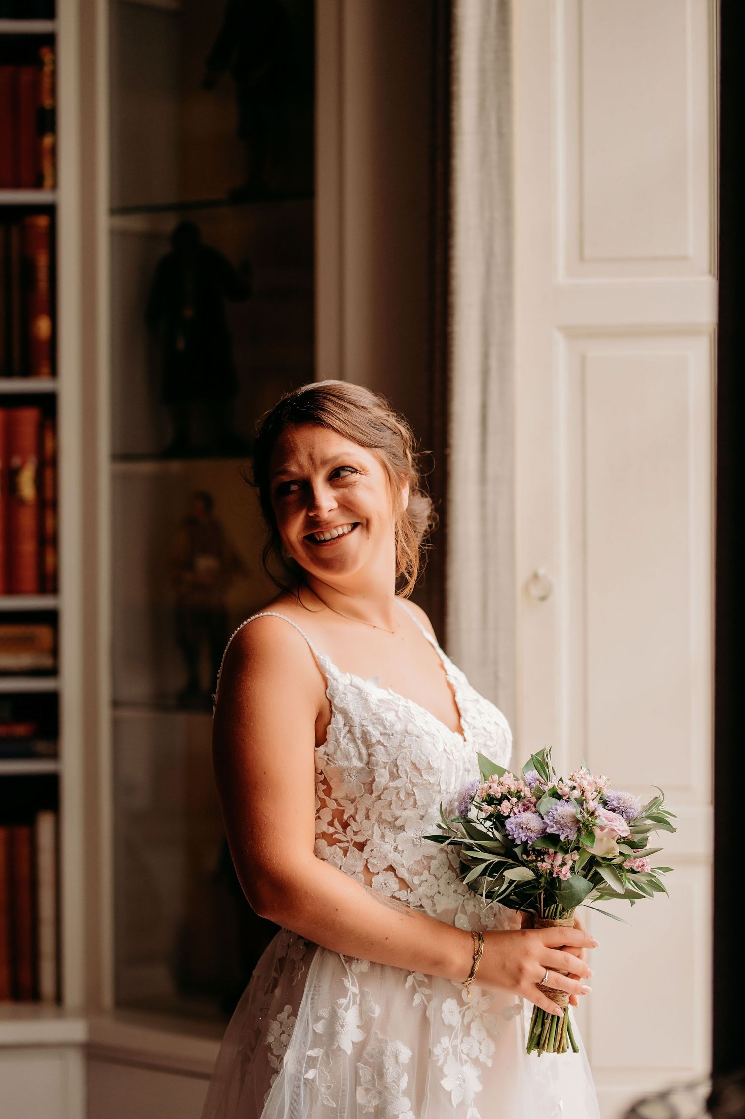 Smiling bride in lace wedding dress holding purple floral bouquet standing in elegant room with bookshelves.