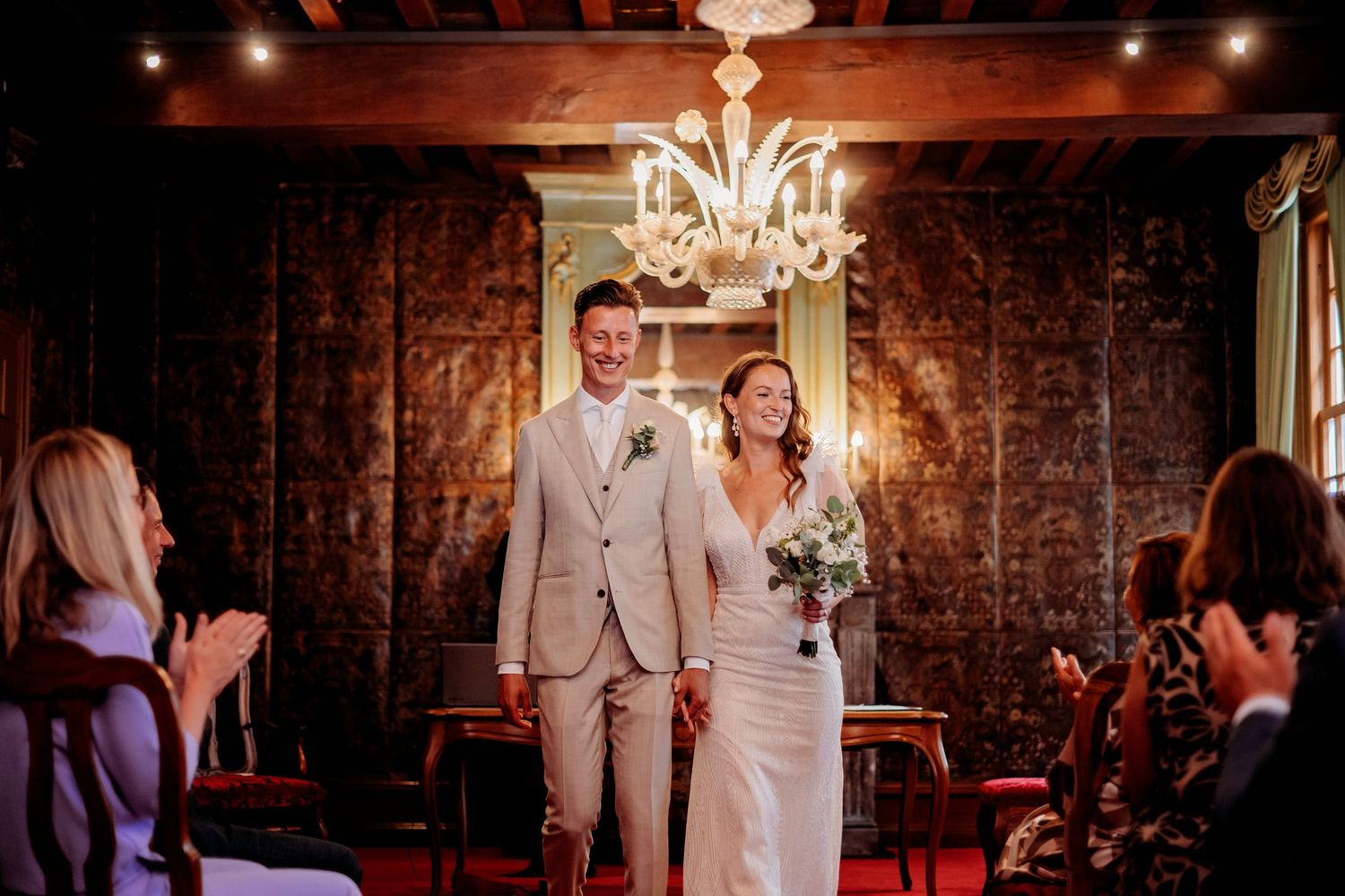 Bride and groom walk down the aisle smiling, guests applauding in elegant chandelier-lit wedding venue.