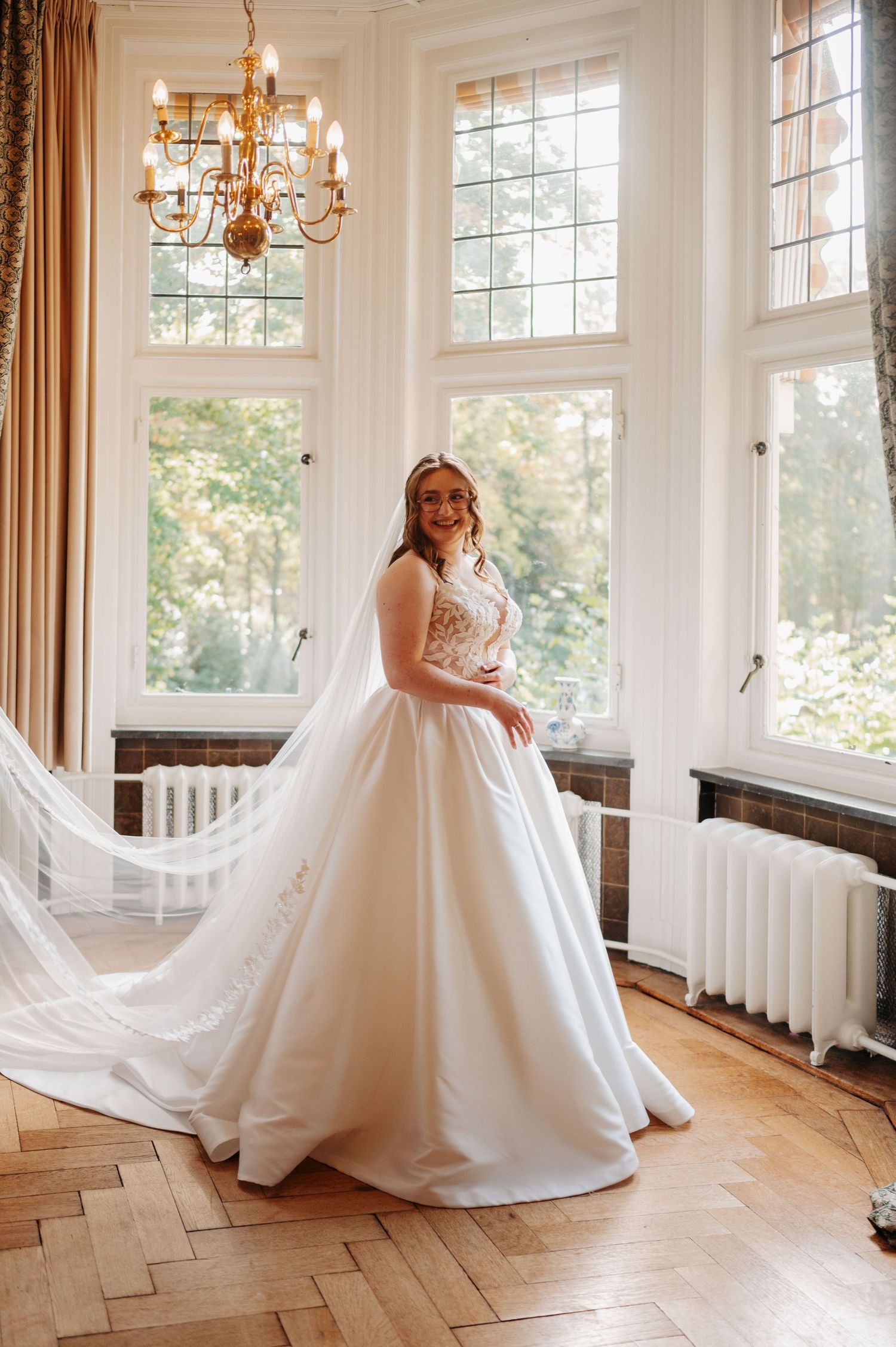 Smiling bride in an elegant ivory satin ball gown with lace bodice and long veil standing in a bright room with tall windows.