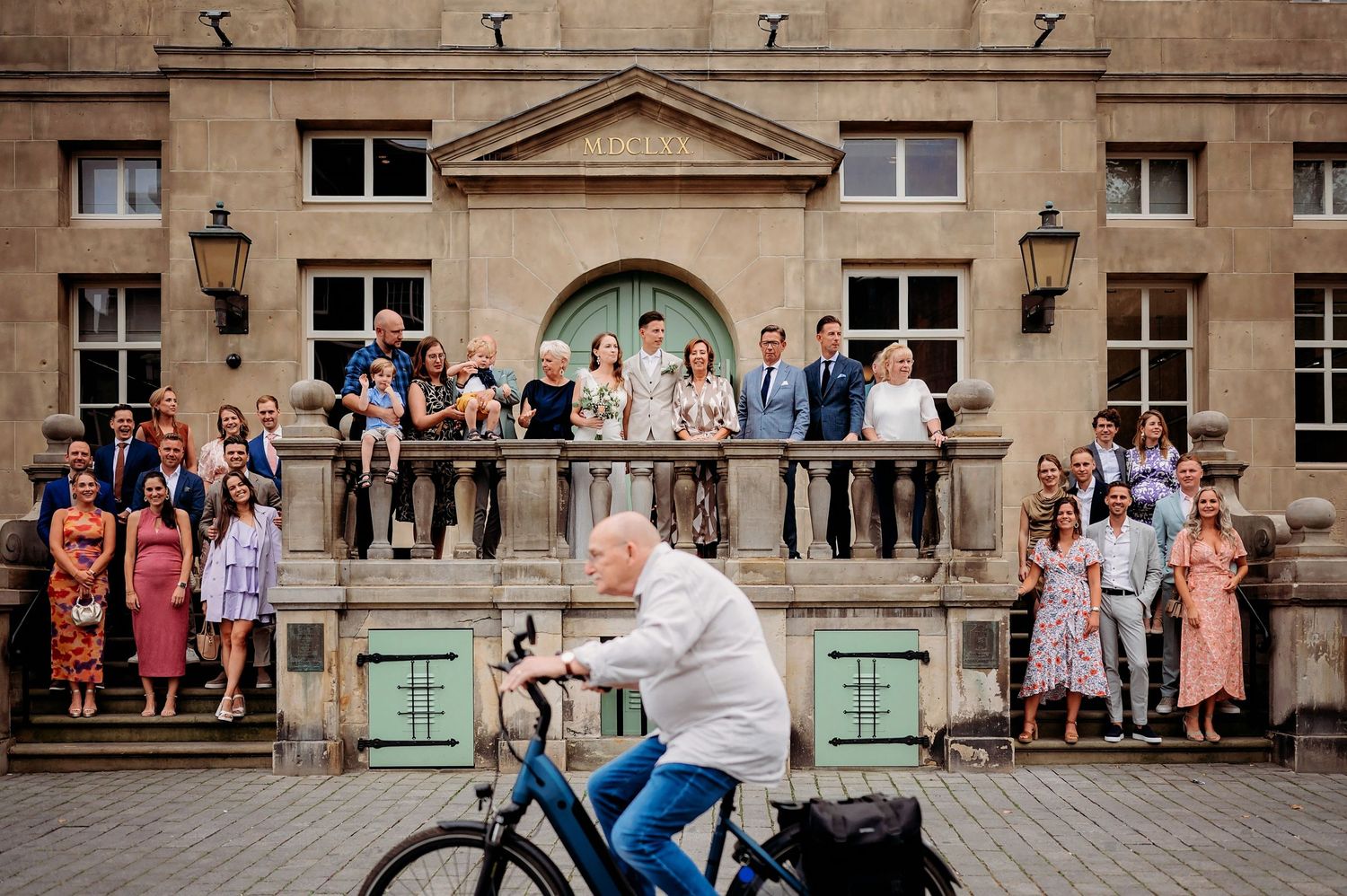 Elderly man cycling past a wedding party posing on a grand building balcony, candid street photography.
