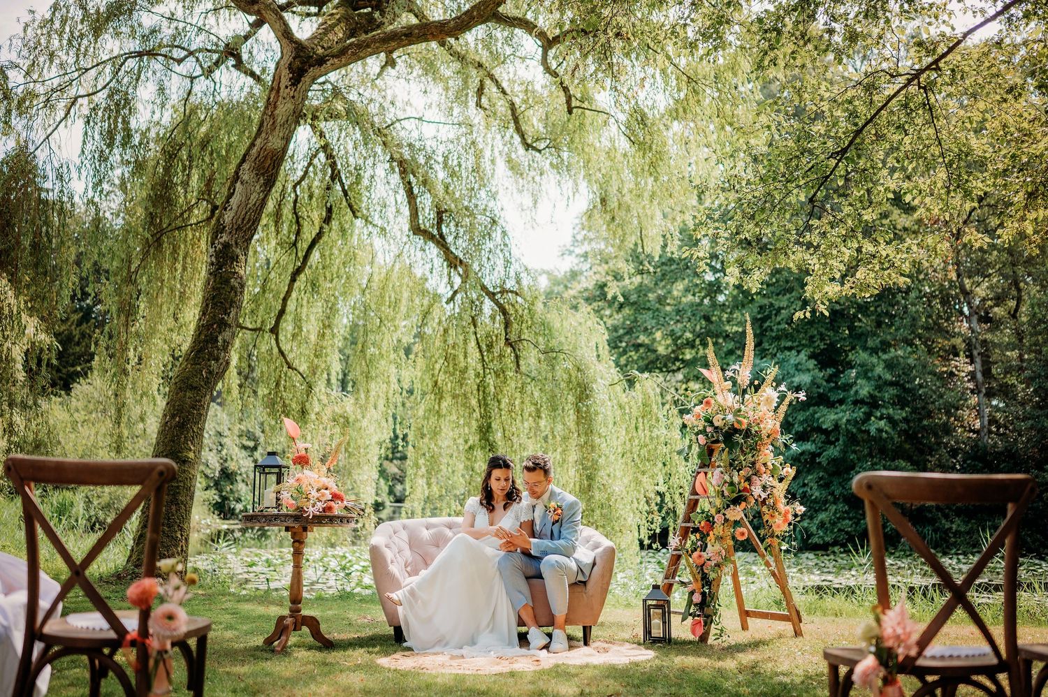 Romantische buitenceremonie onder een wilgenboom met vintage meubels en bloemdecoraties, bruidsfotografie Den Bosch.