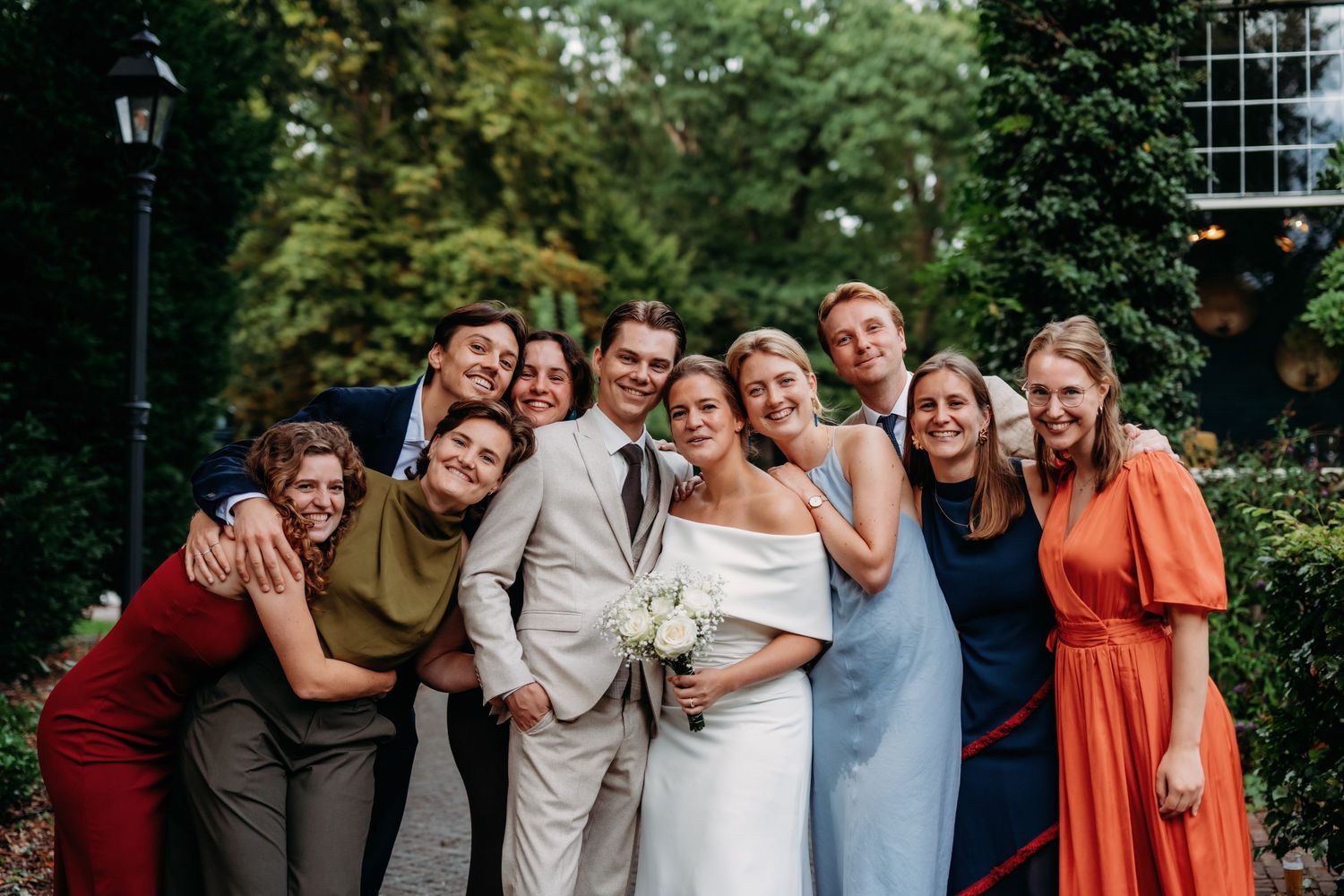 Wedding party group hug with bride and groom surrounded by friends in colorful attire outdoors.