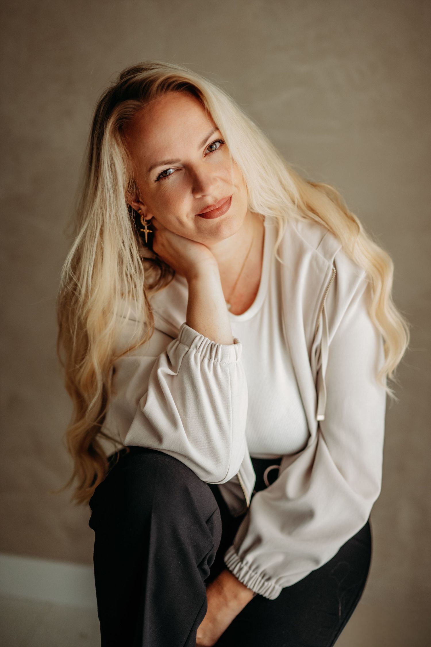 Smiling blonde woman in white blouse and dark pants poses elegantly with hand on cheek against neutral background.