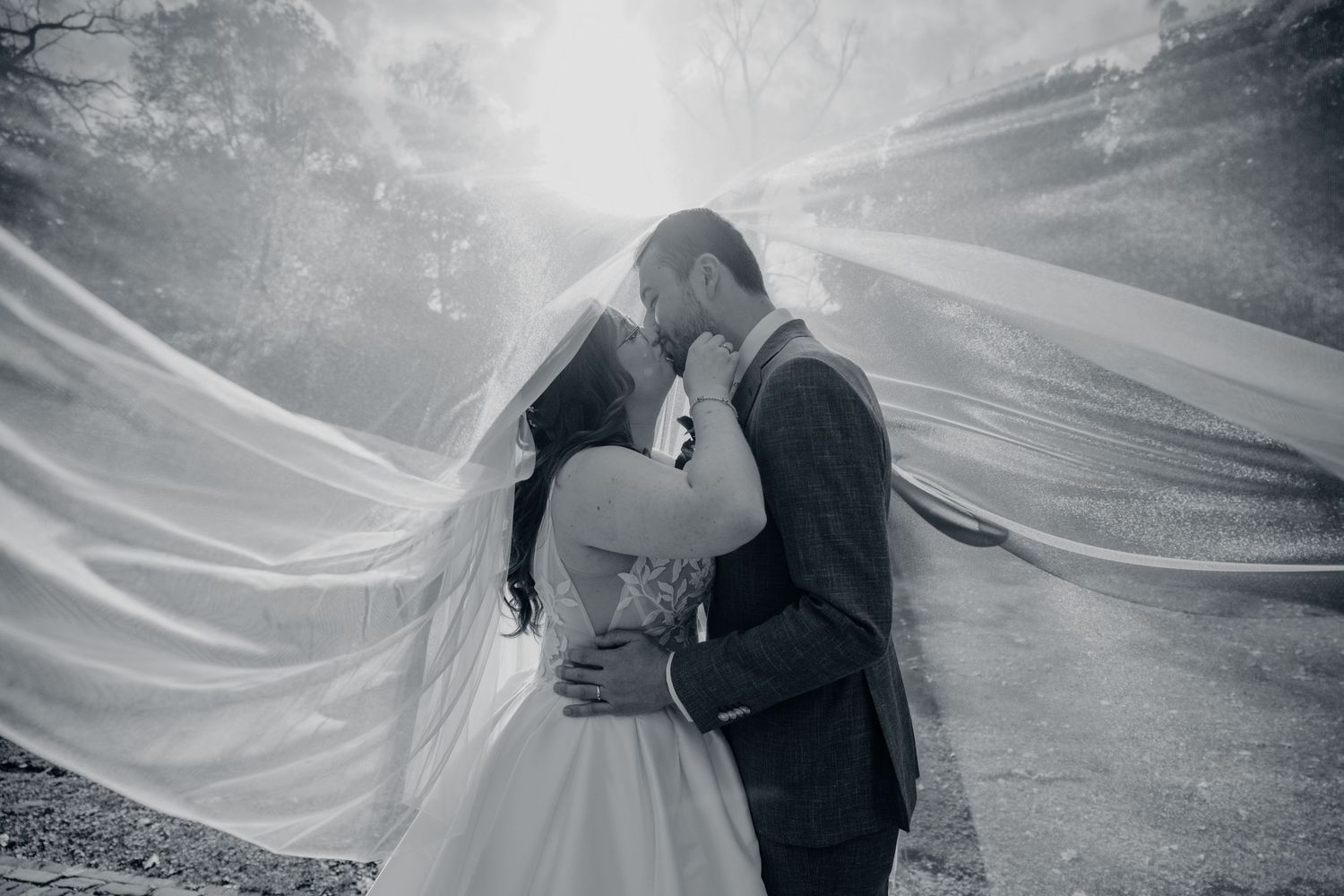 Black and white photo of a bride and groom sharing a kiss beneath a long, flowing wedding veil in a misty, sunlit setting.
