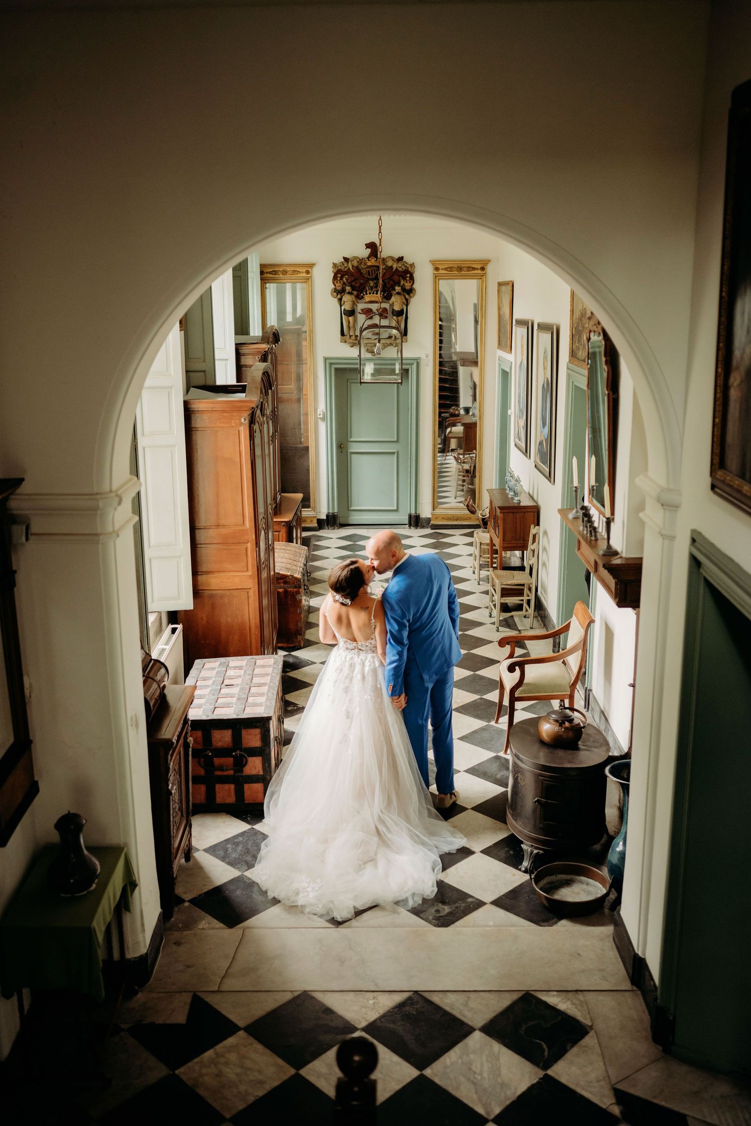 Bride and groom share a romantic kiss under an elegant archway in a grand historic venue with checkered floors.