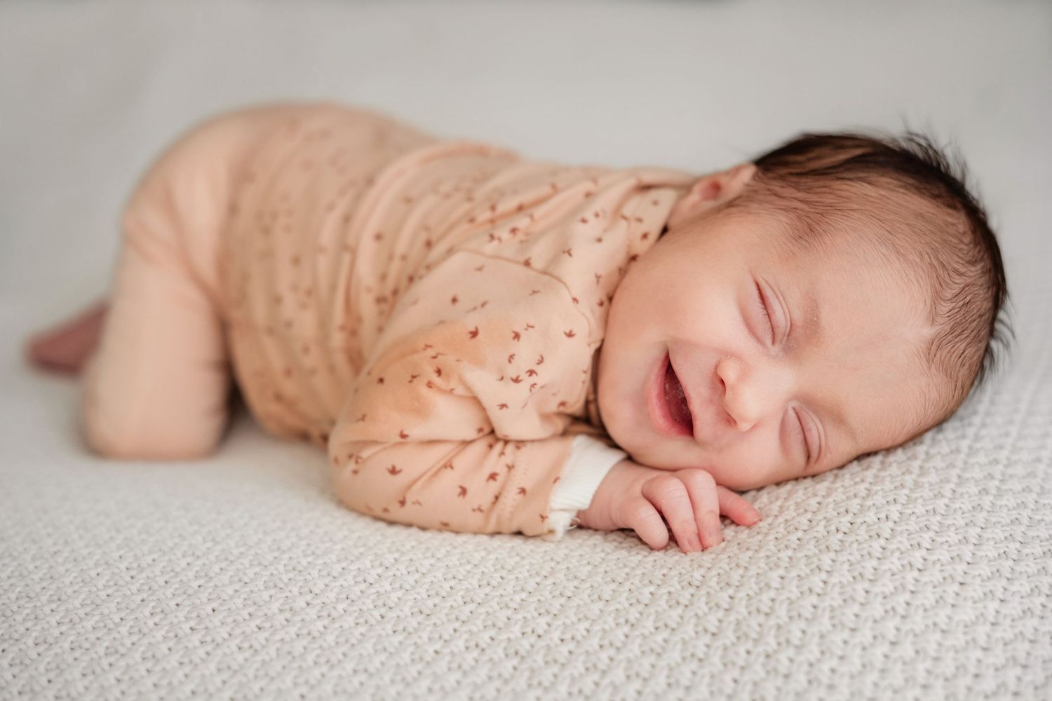 Sleeping newborn in peachy outfit curled up on textured white surface.