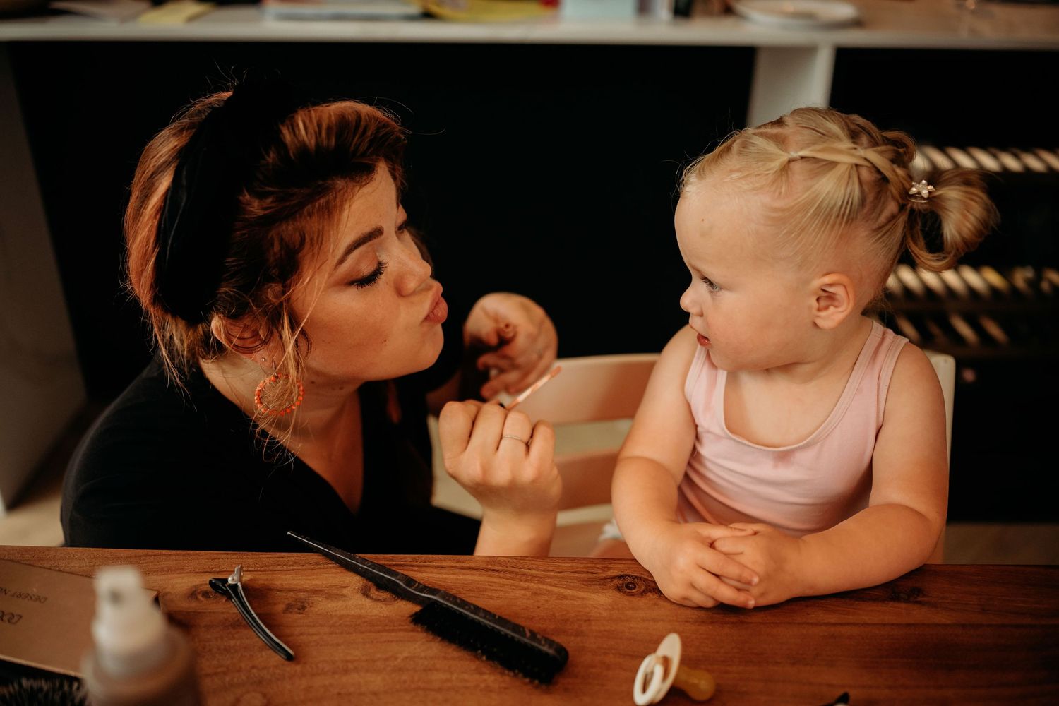 A young woman applies makeup to a toddler girl sitting at a wooden table with beauty tools nearby.