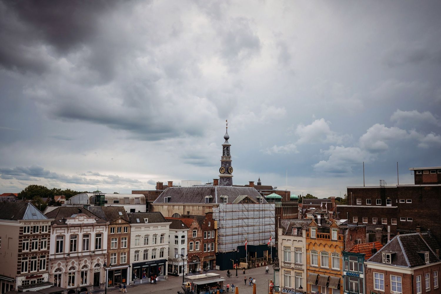 Aerial view of a historic European town square with colorful buildings, a church spire, and dramatic cloudy skies.