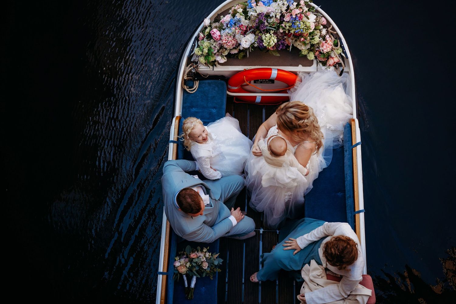 Aerial view of a bride and groom with guests on a decorated boat on dark water, surrounded by floral arrangements.