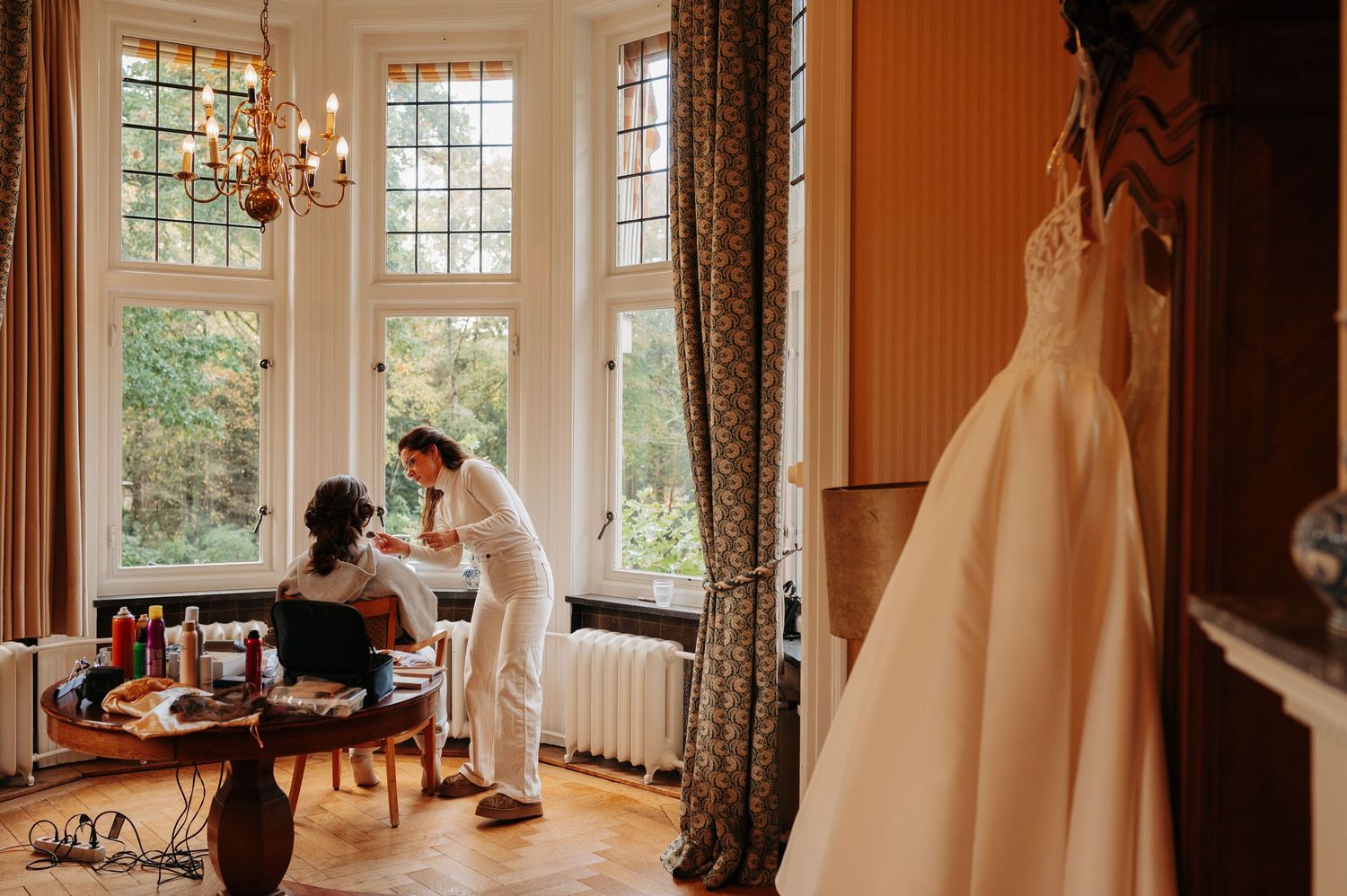 A makeup artist applies bridal makeup to a seated woman in an elegant room with tall windows, while a white wedding dress hangs nearby.