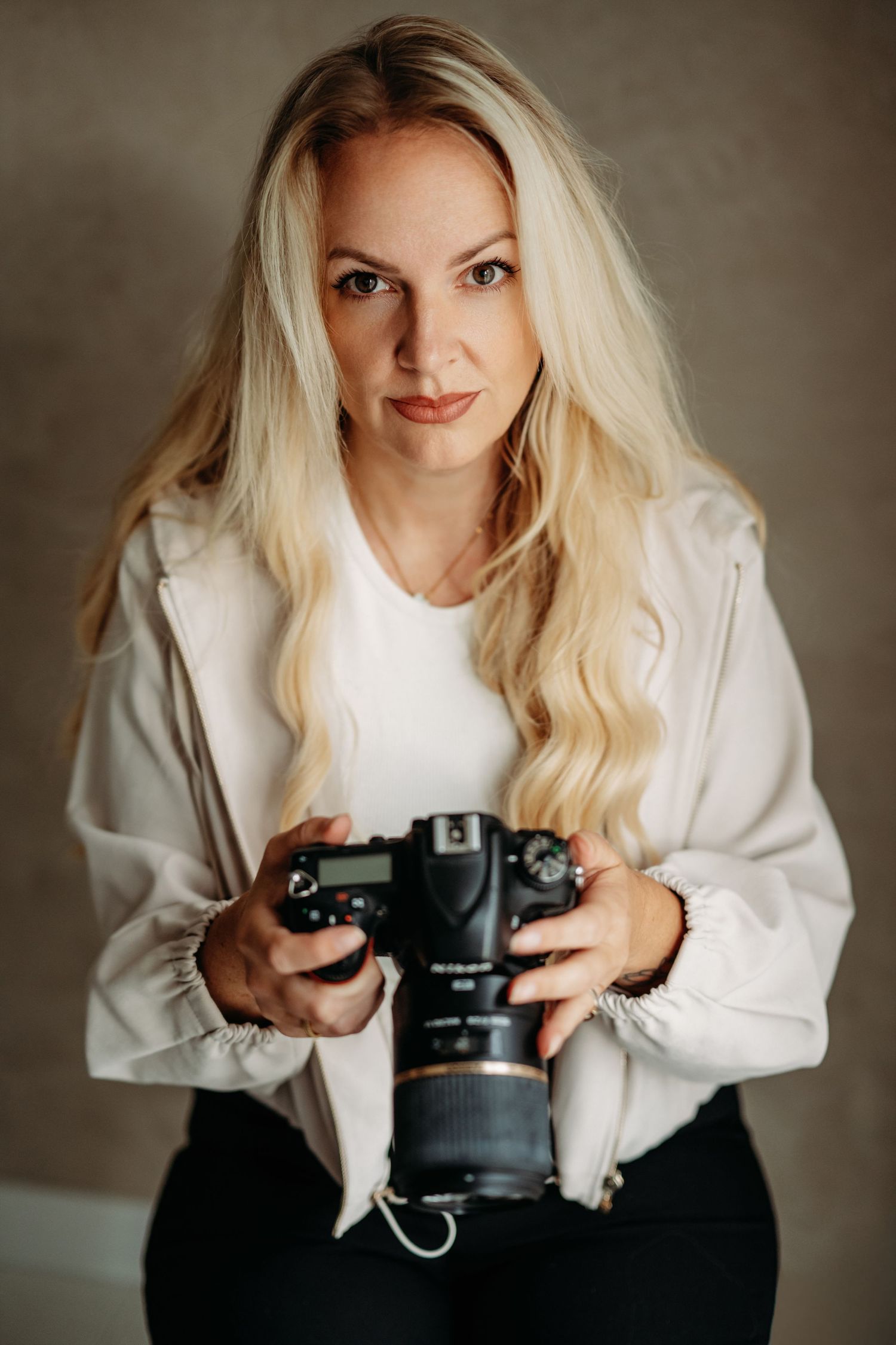 Blonde woman photographer holding a professional DSLR camera, wearing a white jacket, posing in a studio setting.