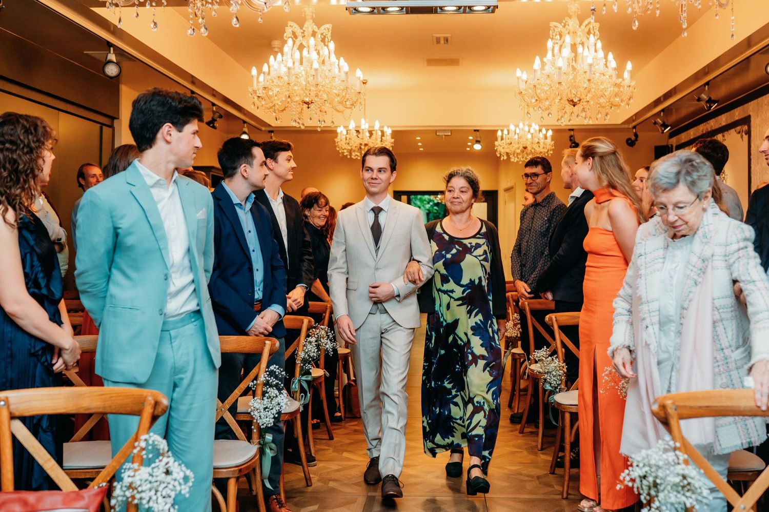 Groom walks down wedding aisle with guest amid chandeliers and wooden chairs.
