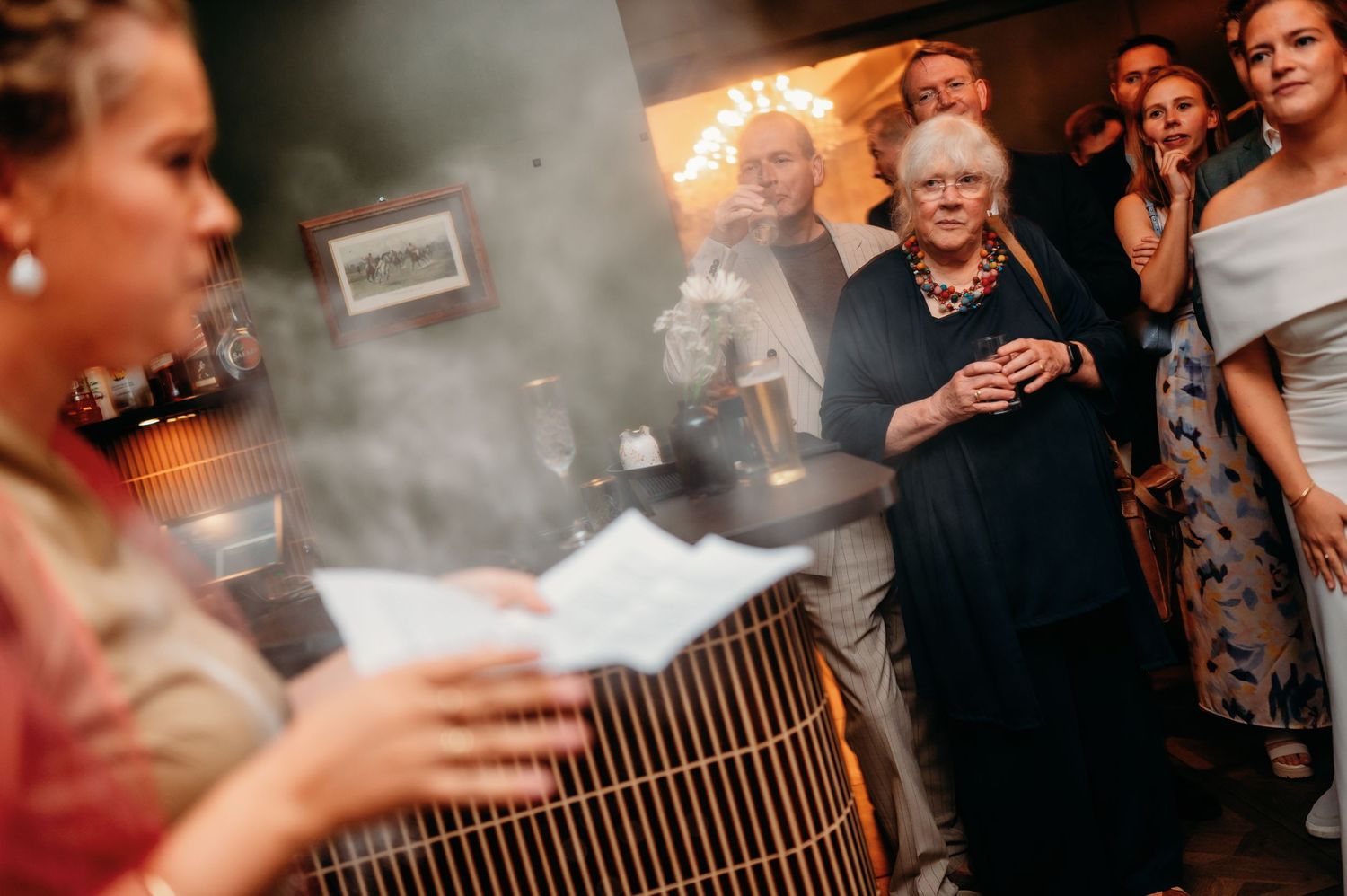 Elderly woman in necklace watches smoke display at indoor evening event with guests in background.
