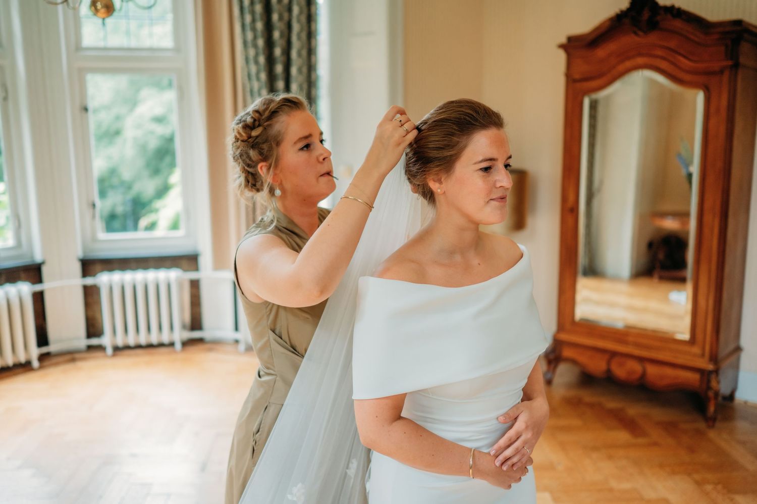 Bridesmaid helping bride with veil in elegant room with antique mirror on wedding day.