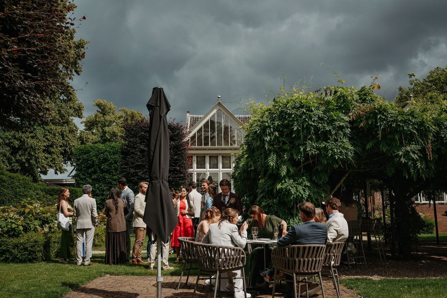 Guests gather outdoors at garden party tables under stormy skies near elegant greenhouse venue.