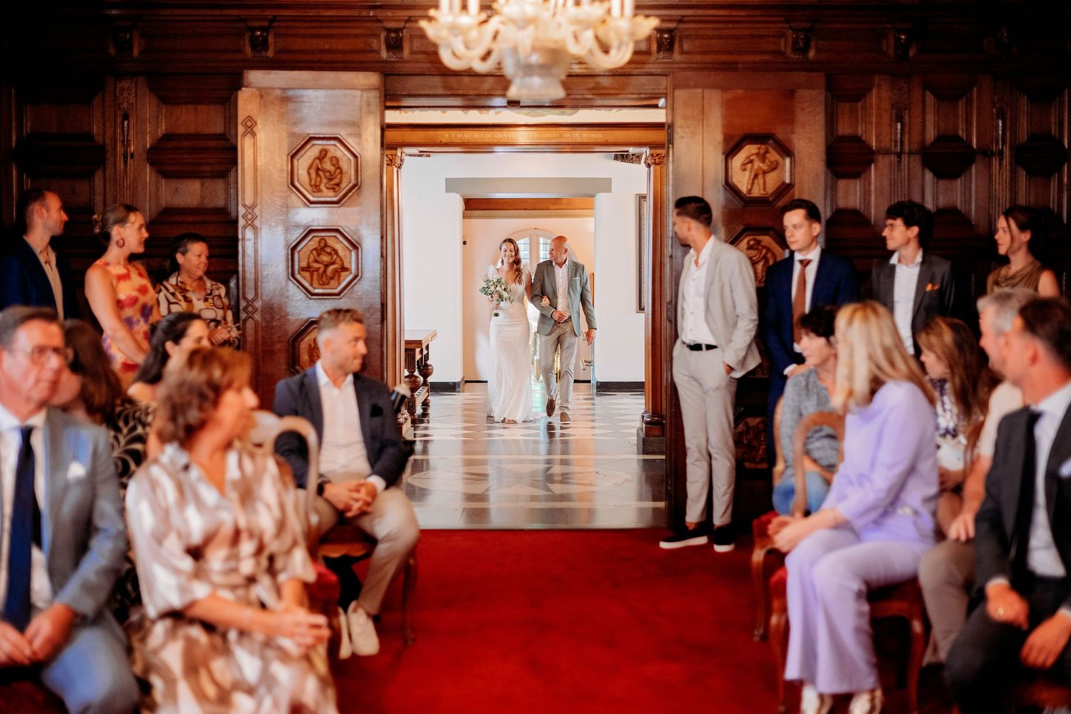 Elegant wedding ceremony in a grand hall with red carpet aisle, guests seated as couple walks toward altar.