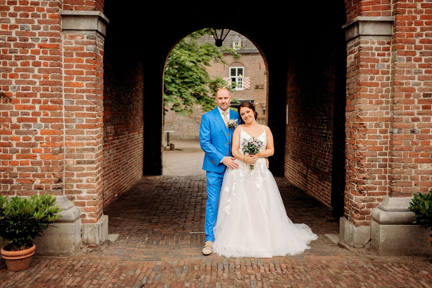Bride and groom pose under brick archway on cobblestone path, groom in blue suit, bride in white gown holding bouquet.