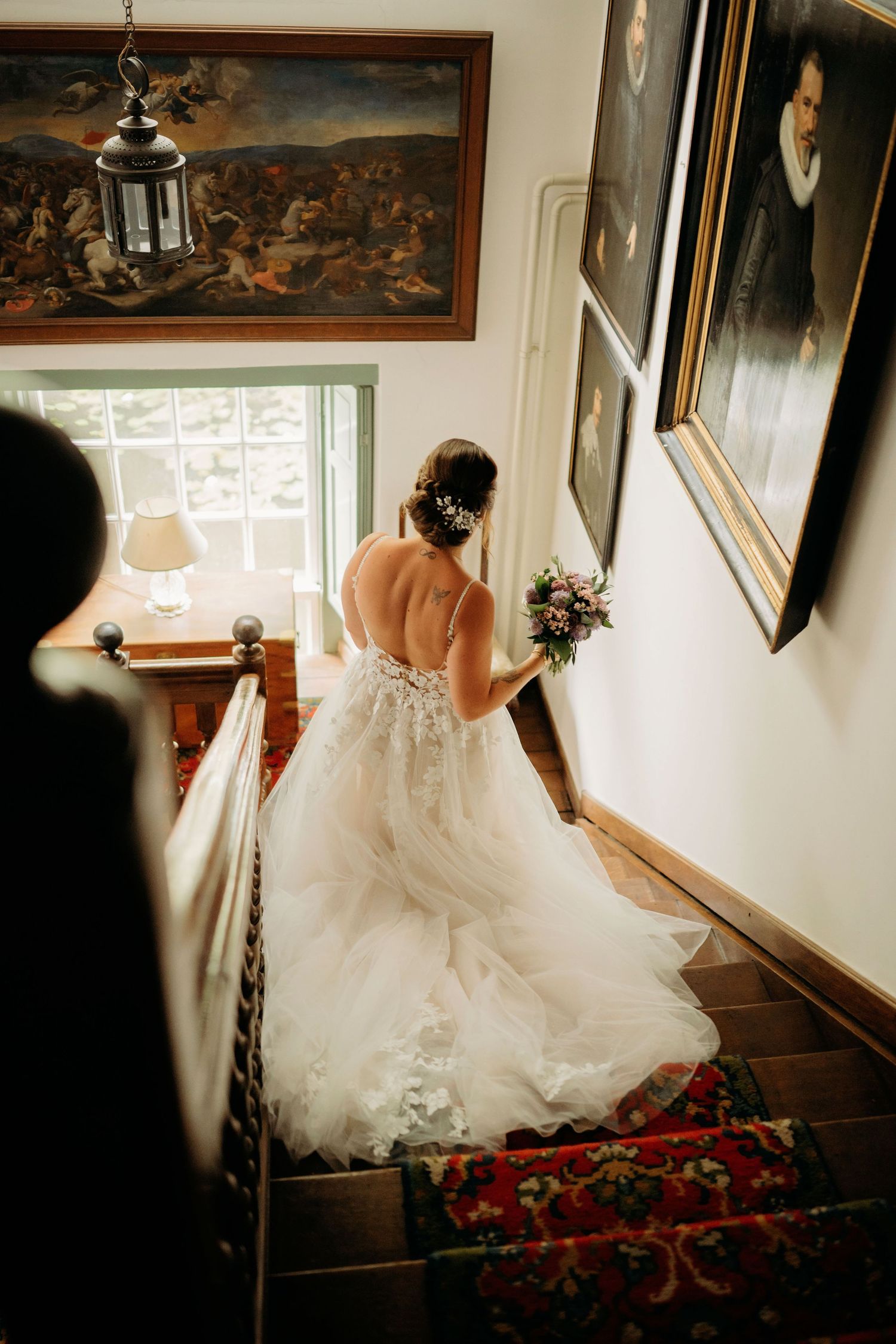 Bride in backless lace wedding gown descending staircase with bouquet past vintage portrait paintings.
