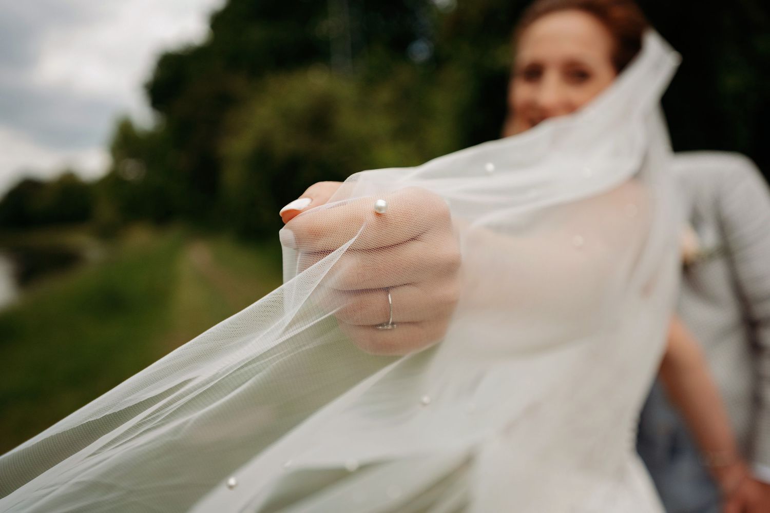 Bride holding flowing white wedding veil outdoors, wearing engagement ring, with greenery in background.
