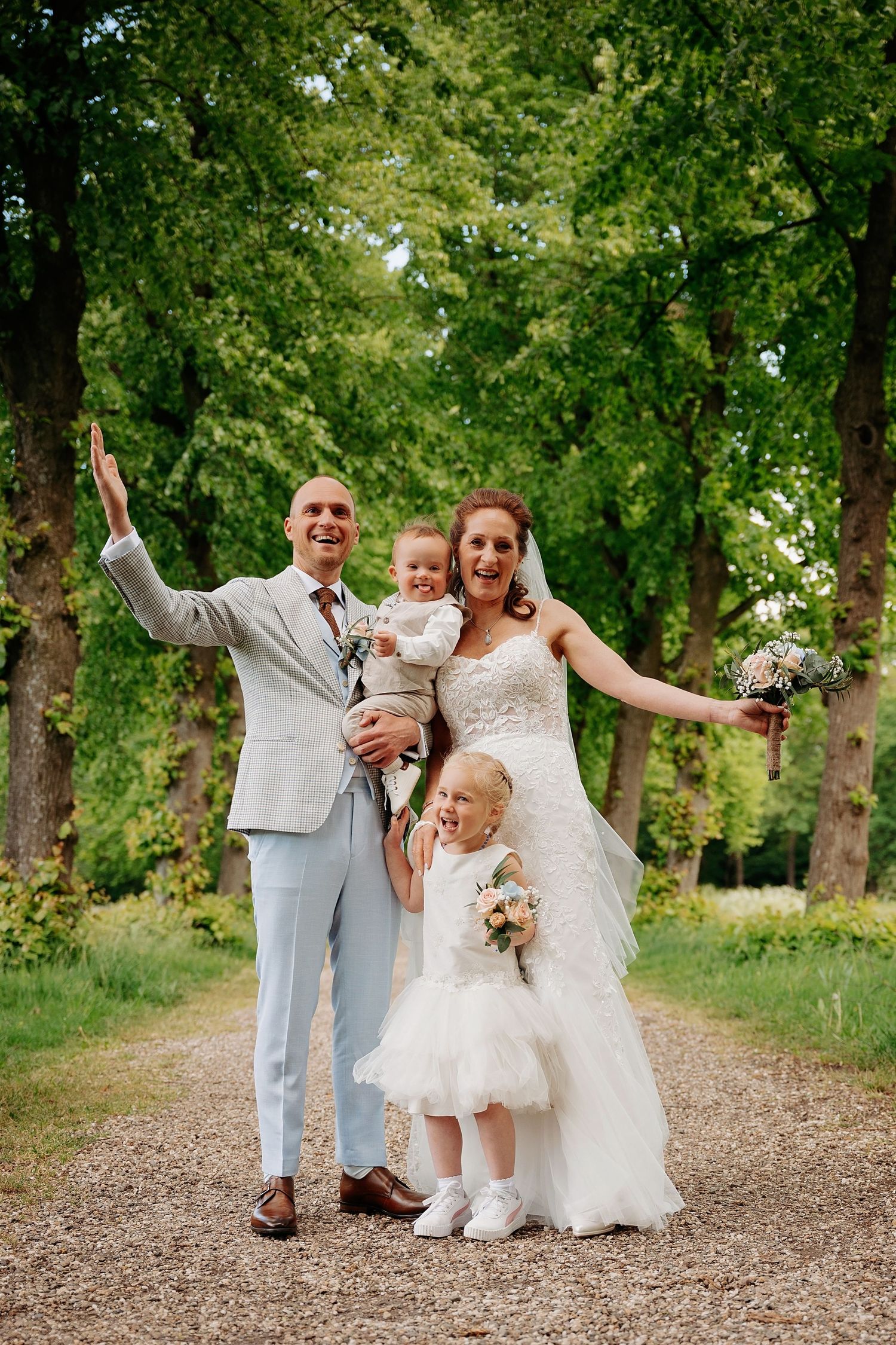Joyful wedding couple with two young children posing on a tree-lined path, bride holding bouquet in white gown.