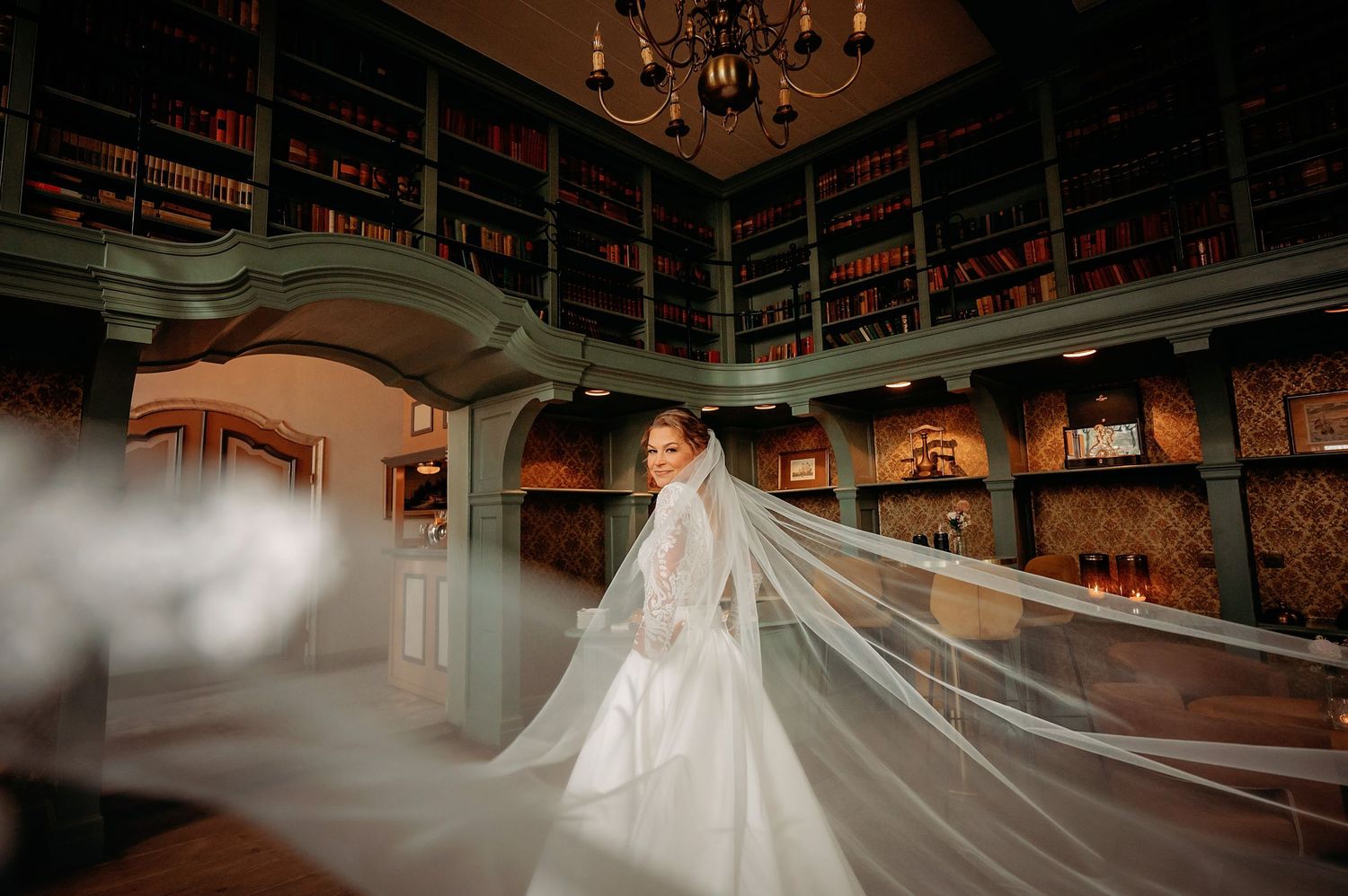 Vloeiende witte jurk en sluier gefotografeerd in elegante bibliotheek met sierlijke architectuur en boekenplanken, bruidsfotografie Brabant