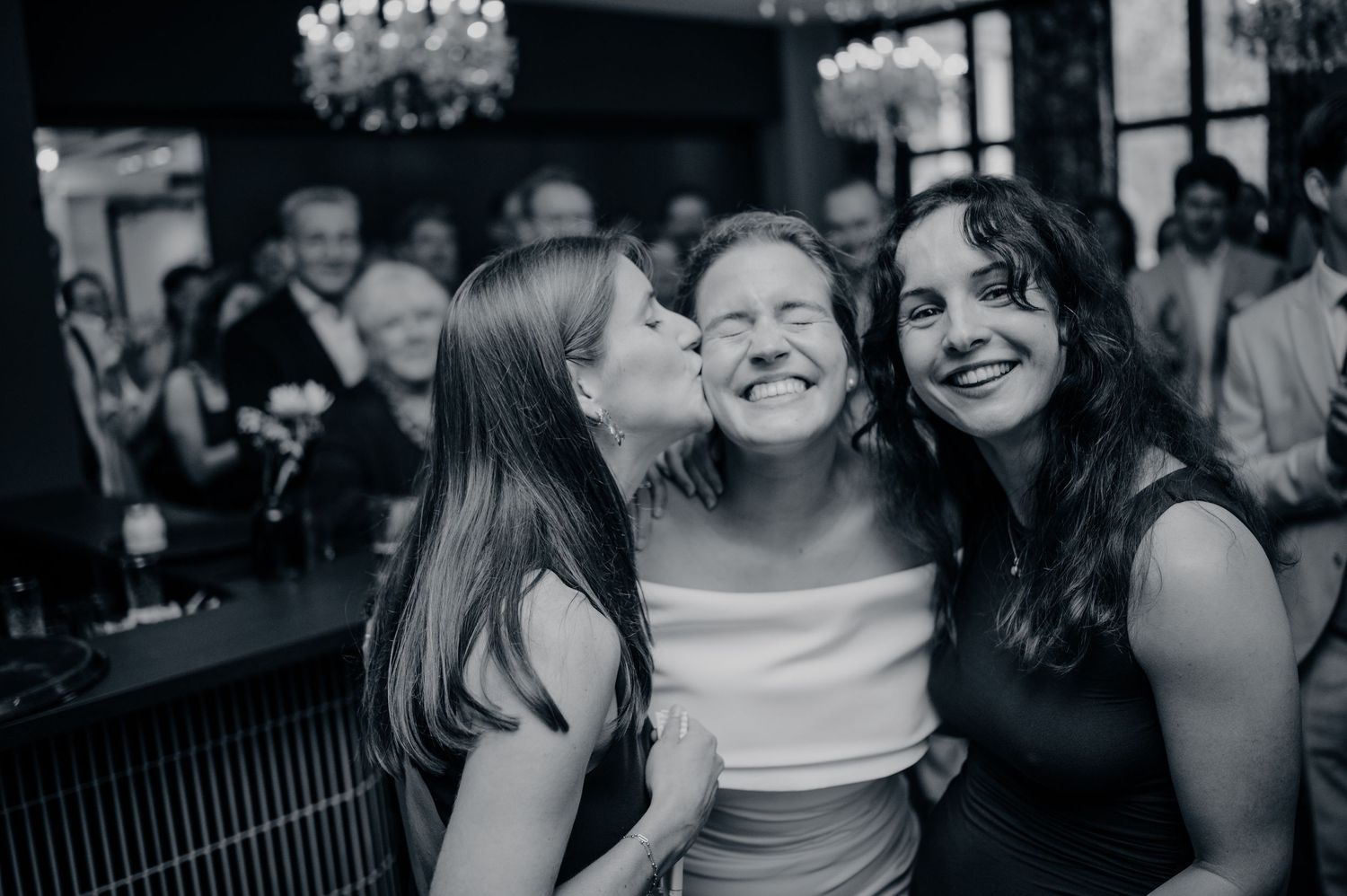 Black and white photo of bride being kissed on the cheek by two smiling friends at a wedding celebration.