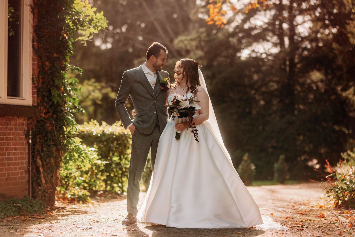 Bride in a white gown holding a bouquet and groom in a gray suit embracing outdoors in warm autumn sunlight near a brick building.