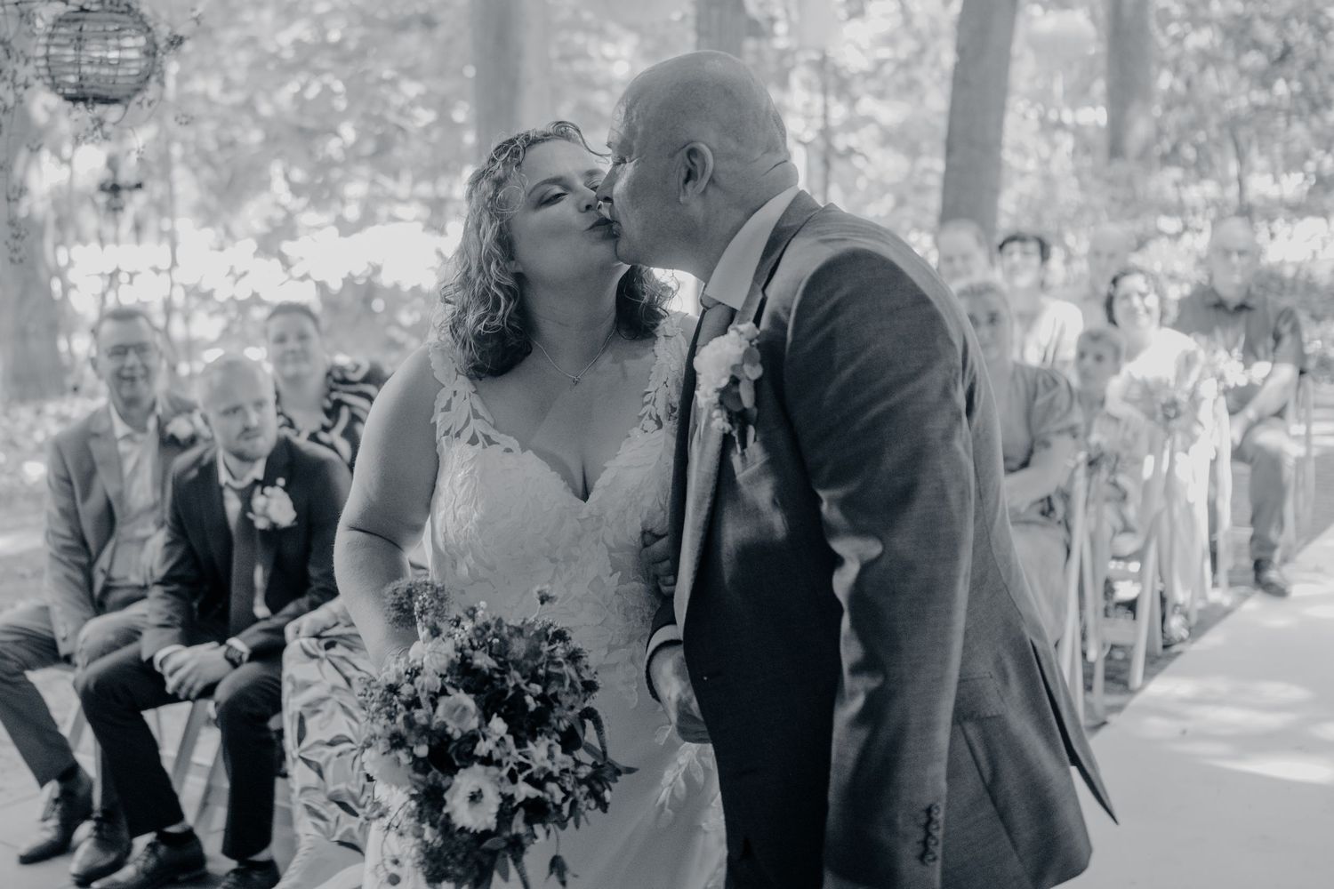 Black and white photo of a bride and groom sharing a kiss at their outdoor wedding ceremony with guests seated nearby.
