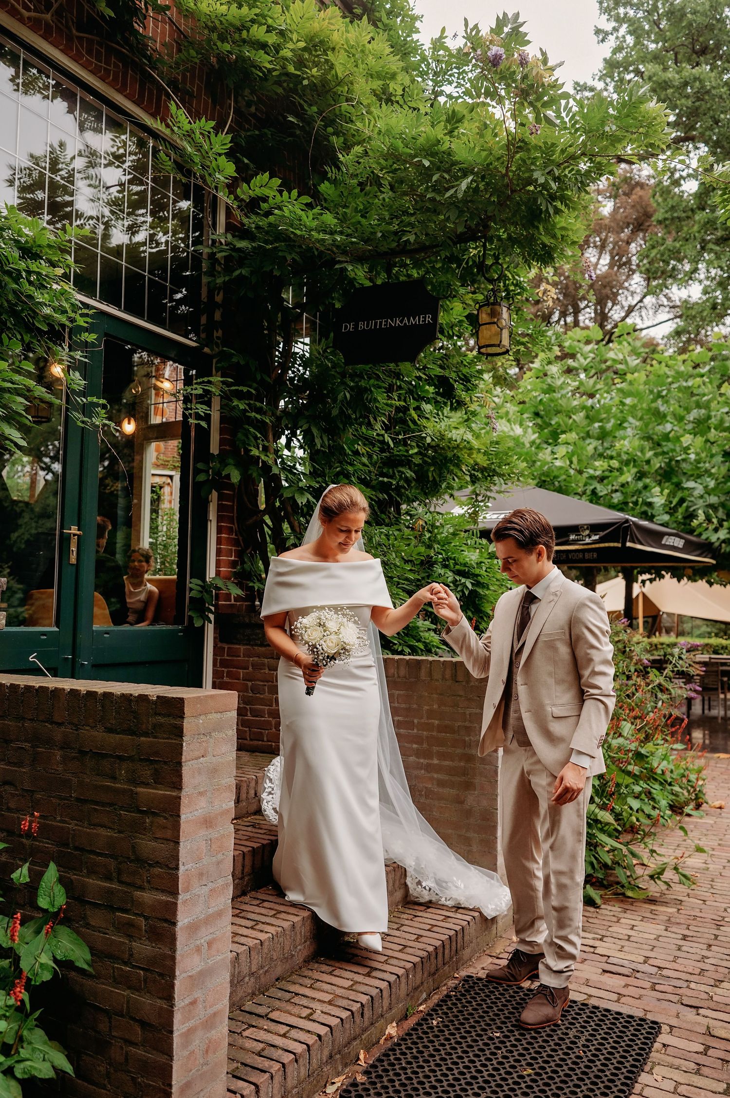 Groom helps bride descend brick steps outside a green ivy-covered venue while she holds a white bouquet.