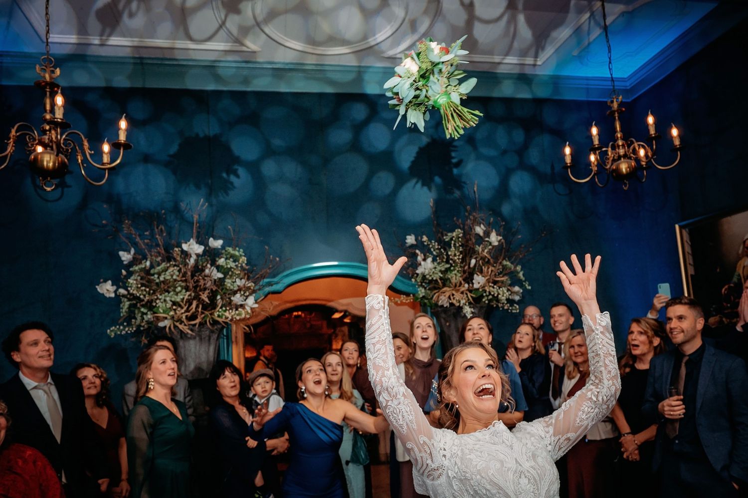Bruiloftsgasten dansen met opgeheven armen onder kroonluchters, spontane bruidsfotografie bij Kasteel Wijenburg Echteld, Brabant.