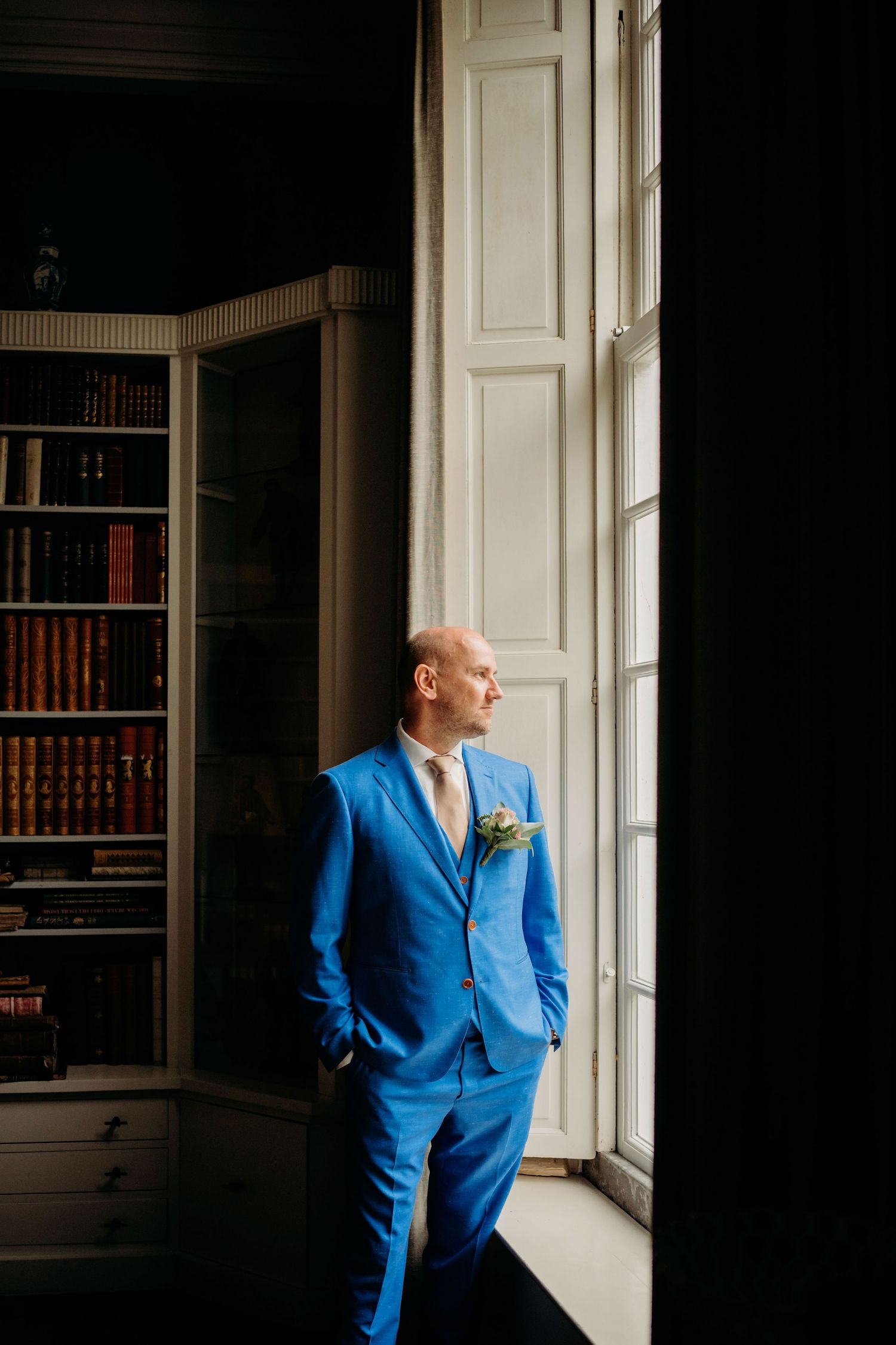 Groom in a bold blue suit standing by a tall window in an elegant library, looking thoughtfully outside.