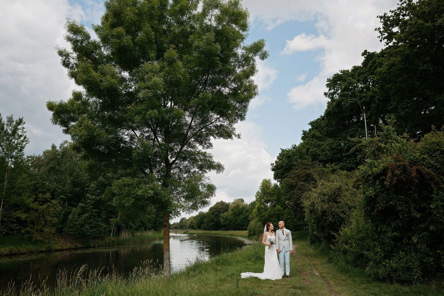 Bride and groom standing together on a lush green path beside a tranquil river surrounded by tall trees.