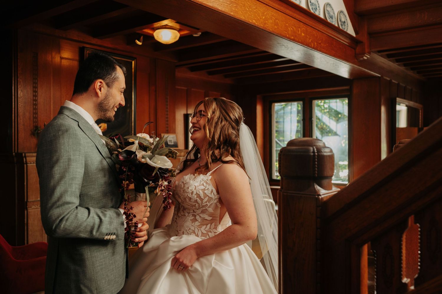 Bride in a lace gown and veil holding a bouquet smiles at her groom in a gray suit inside a rustic wood-paneled lodge.