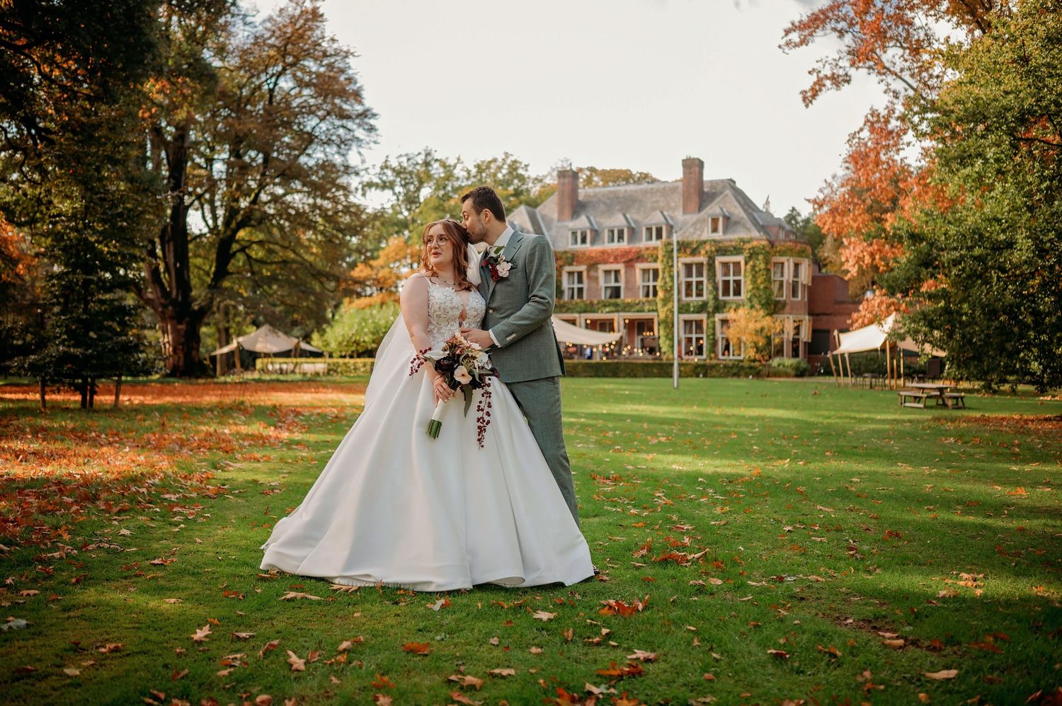 Bride in a white gown and groom in a gray suit embrace on a sunlit autumn lawn in front of a historic brick manor house.