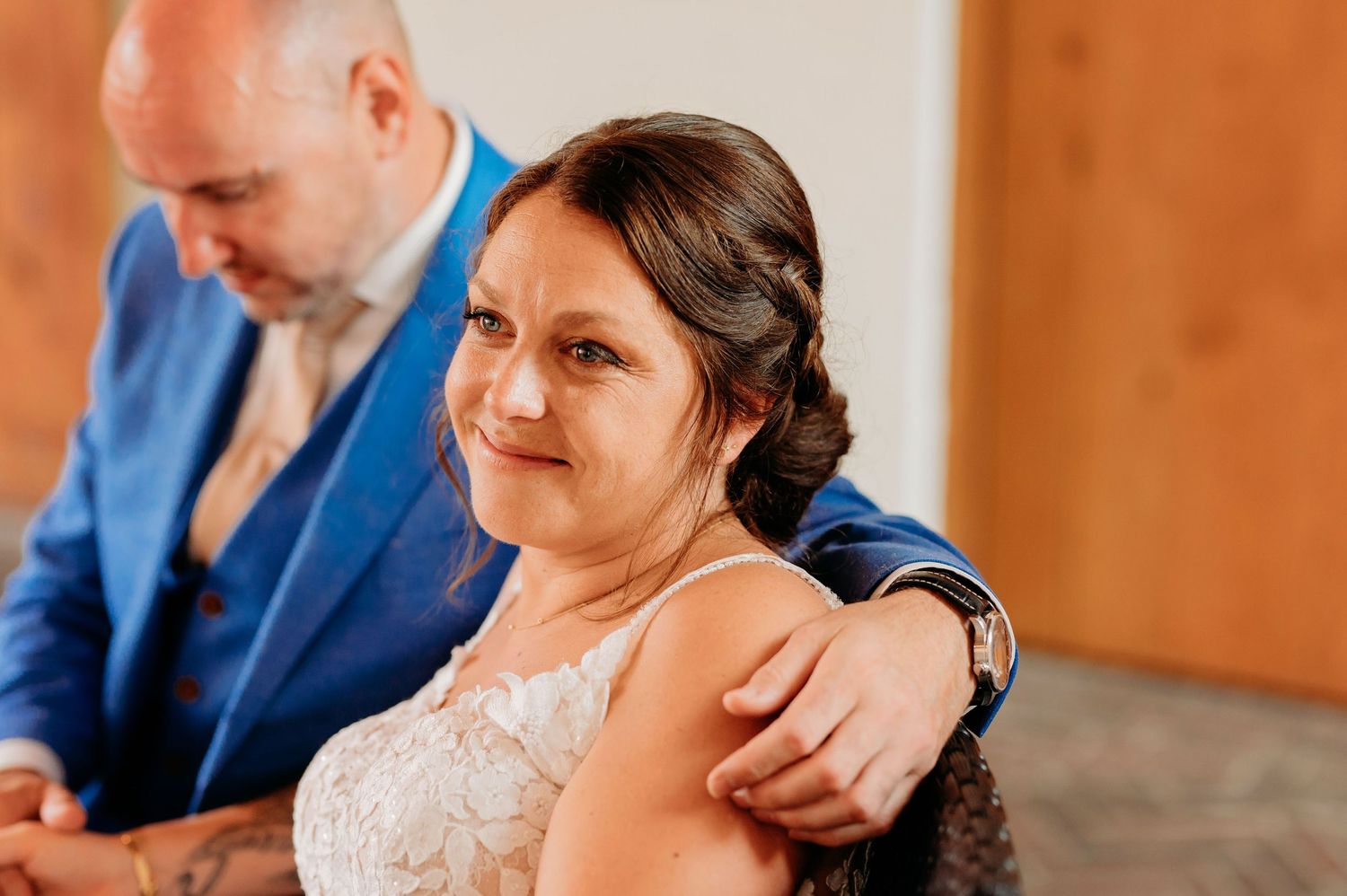 Emotional bride in lace wedding dress with groom in blue suit placing hand on her shoulder.